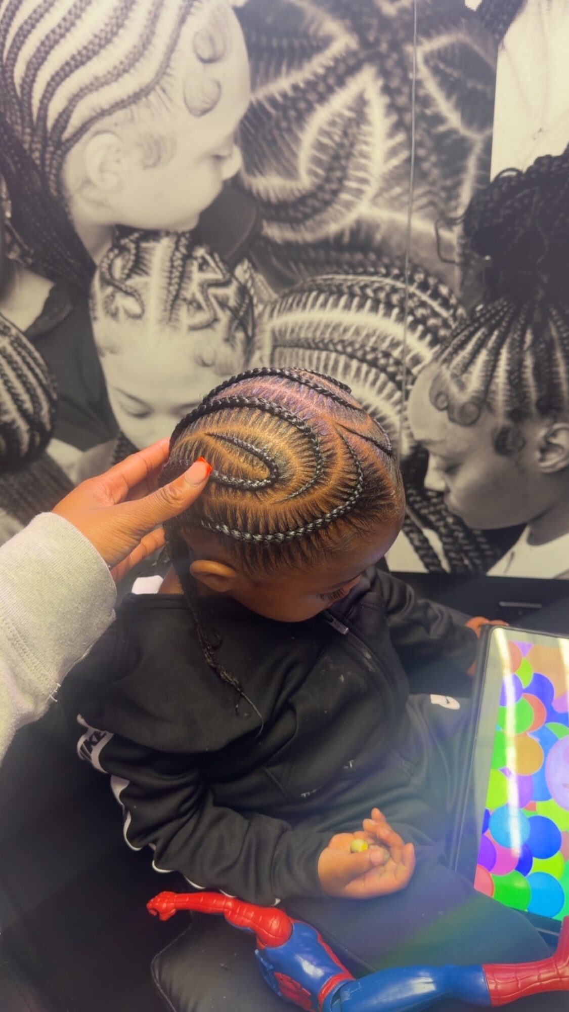 Child with braided hair looking at a colorful tablet, surrounded by black-and-white images of children with braided hair, in a museum or exhibit.