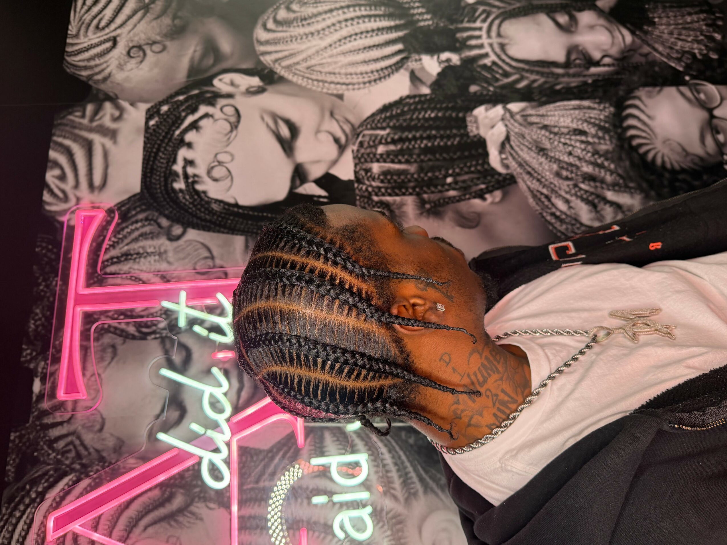 Person with braided hair and a white shirt, holding a phone in front of a black-and-white mural of faces, neon sign in background