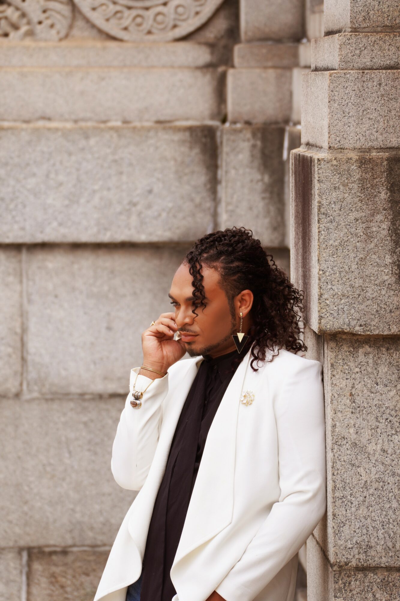 Woman in white blazer and black top leaning against stone wall, touching her face, outdoors.