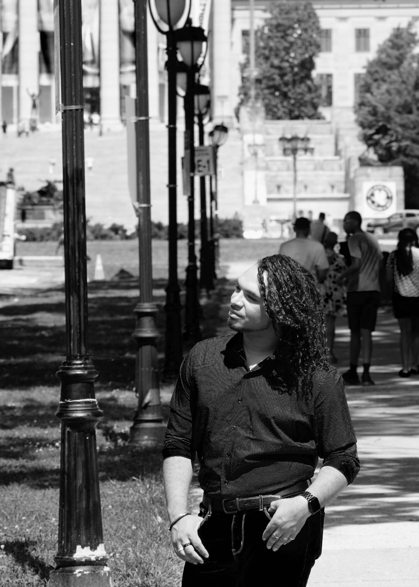 Woman with curly hair standing outdoors near street lamps, with people walking in the background.