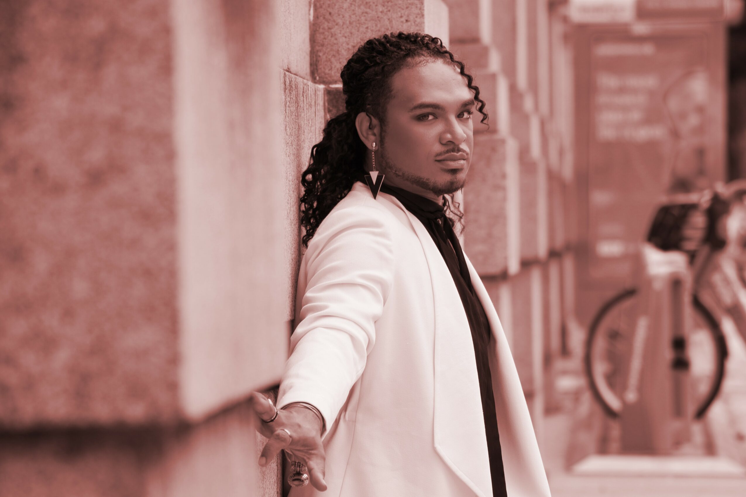 Person with long hair leaning against a wall, wearing a white jacket, in an urban setting.