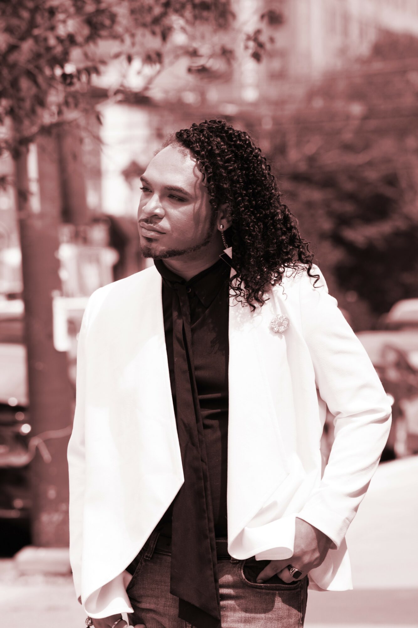 Woman with curly hair in a white blazer and black shirt outdoors, hands in pockets, looking to the side.