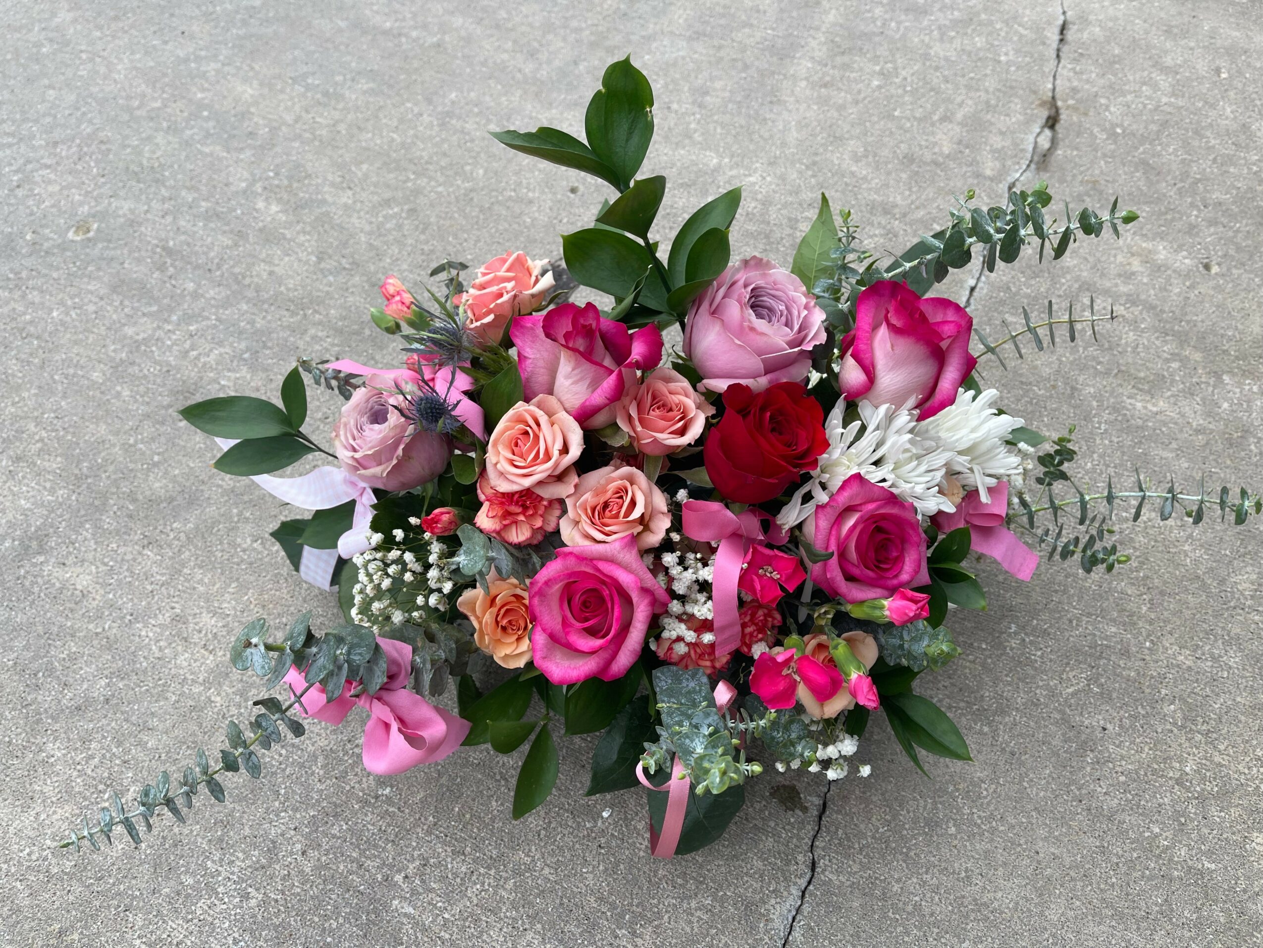 Colorful bouquet of pink, red, and white roses with green leaves and small white filler flowers on a gray surface.