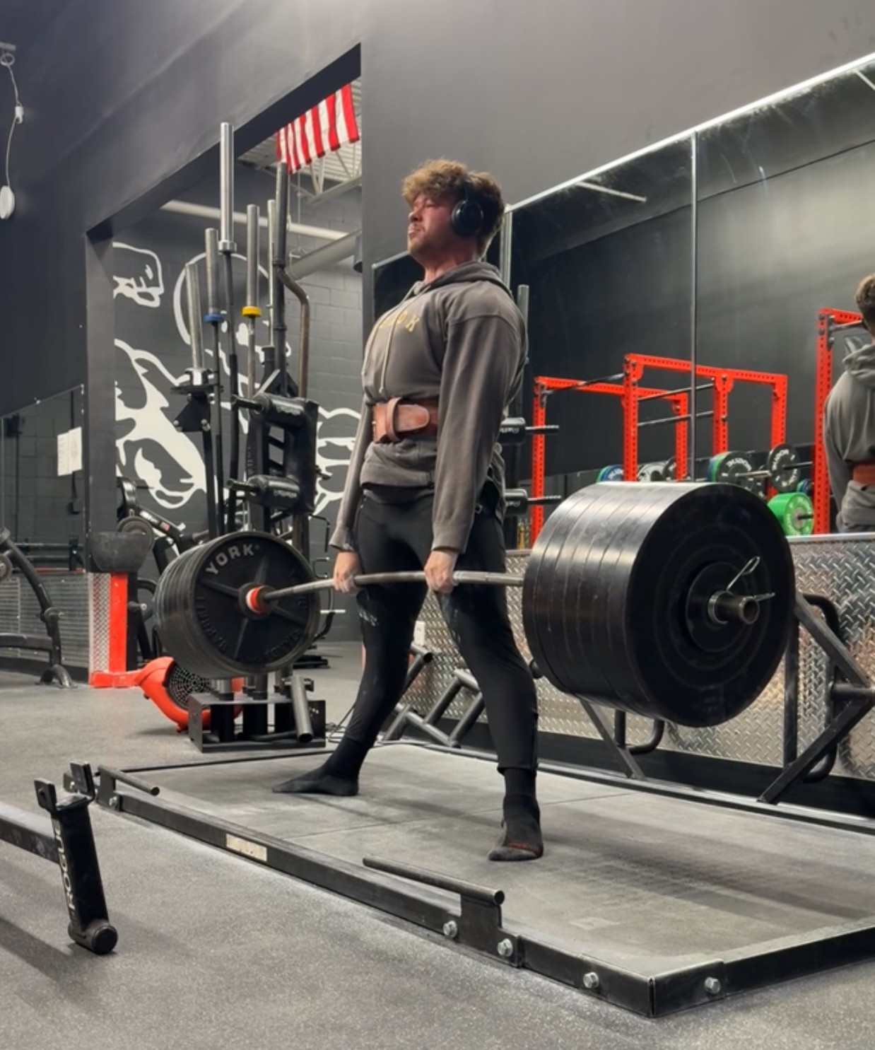 Person performing a deadlift with a loaded barbell in a gym, surrounded by equipment and black walls.