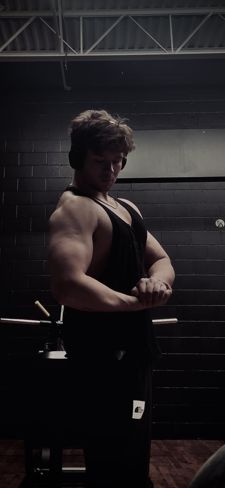 Person with muscular arms and curly hair standing with arms crossed in a gym, dark background, overhead lighting.