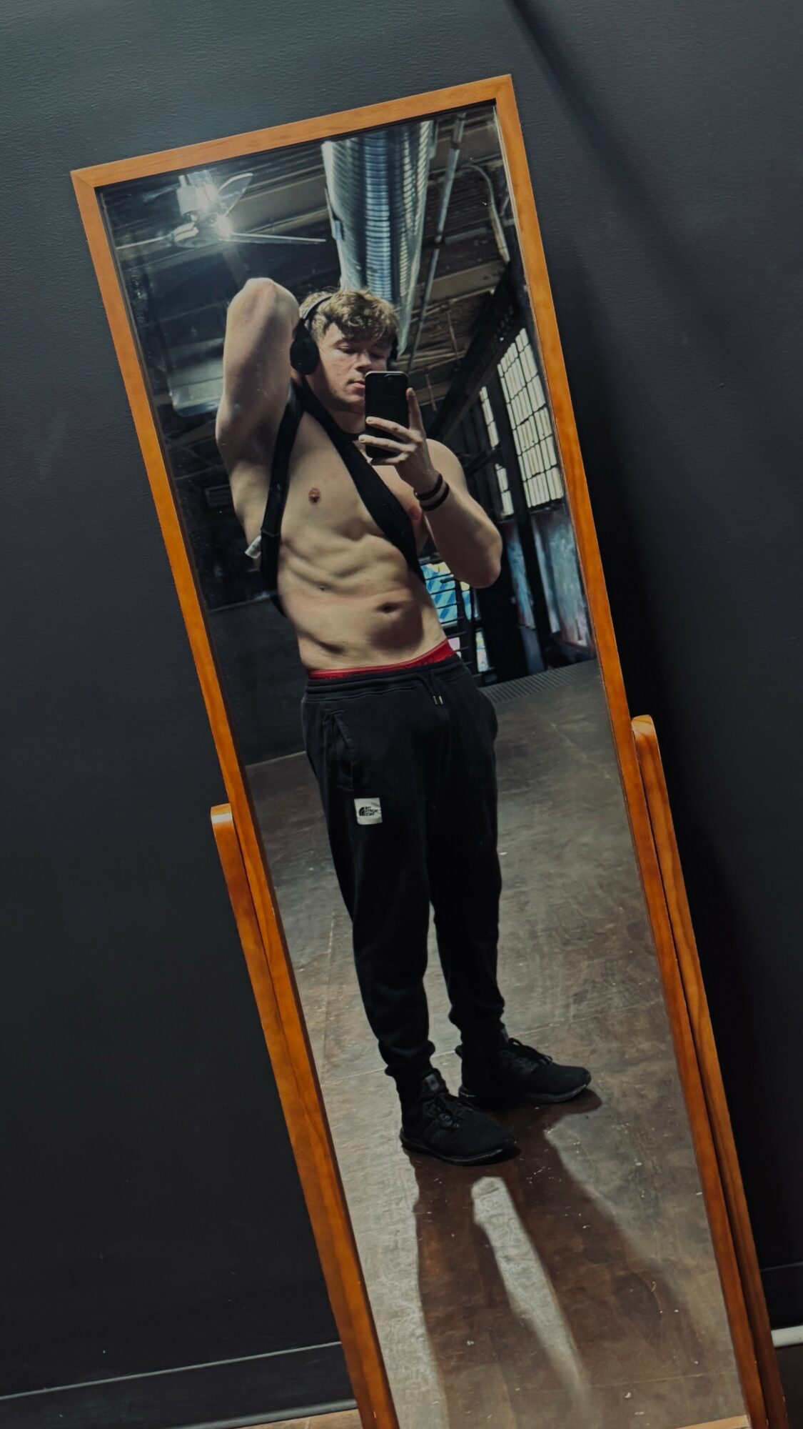 Young man taking a mirror selfie, shirtless, in a room with a wooden frame mirror, wearing black pants and sneakers.