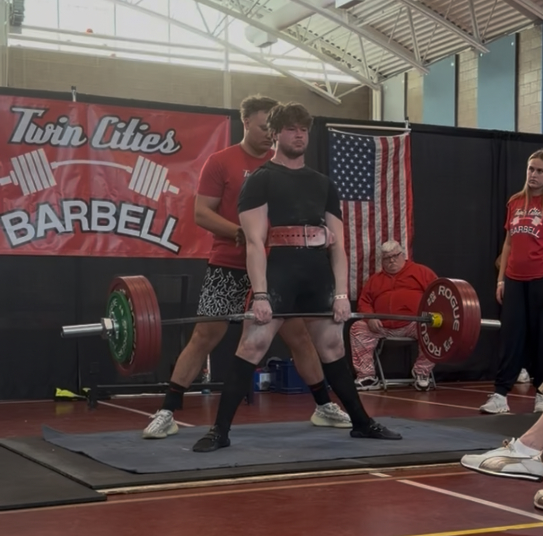 Person in black lifting a barbell with red and green weights, two people behind, and a banner reading 'Twin Cities Barbell'.