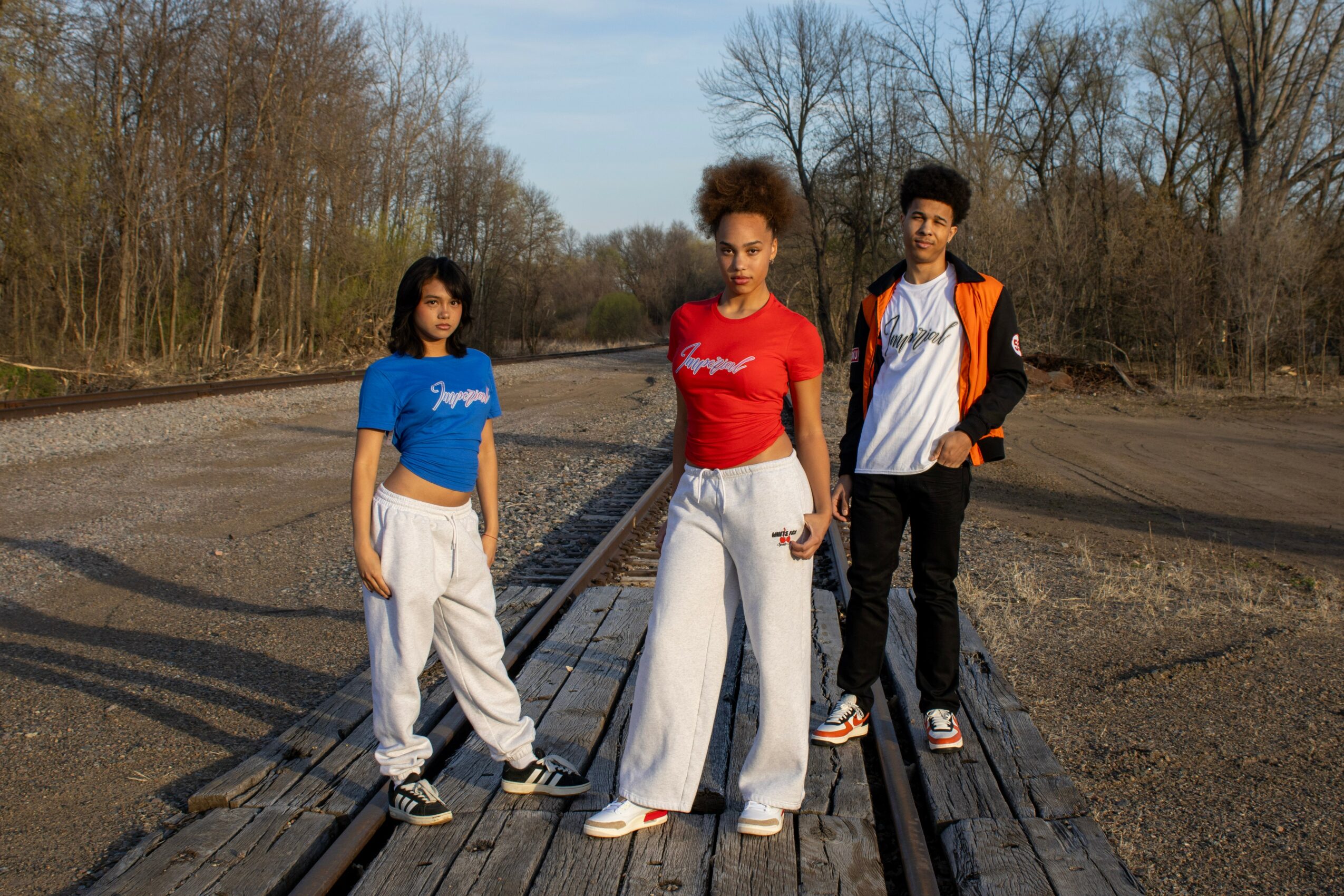 Three young people standing on a wooden pathway outdoors with trees in the background, wearing casual clothing.