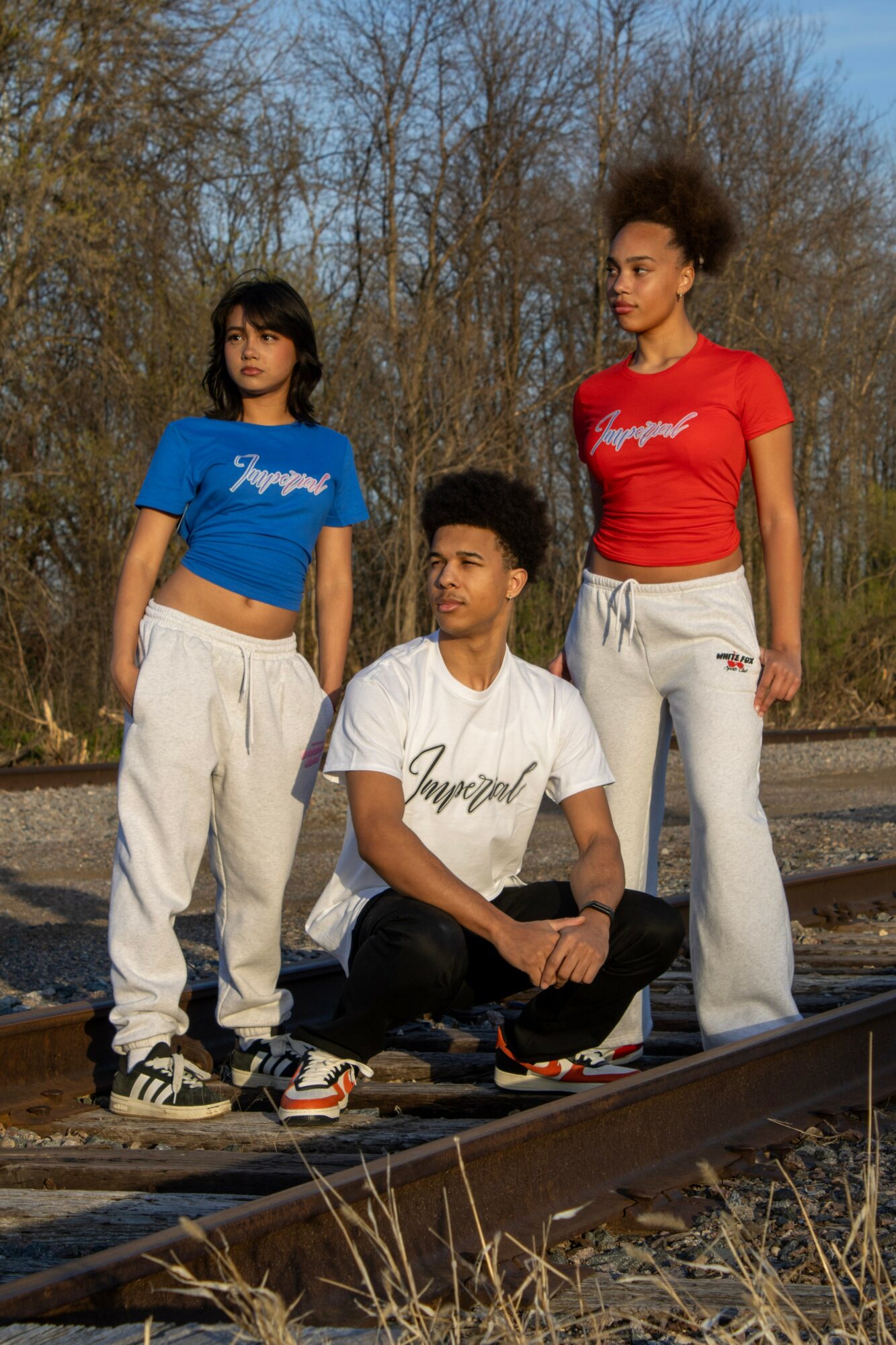 Three young people standing on train tracks outdoors, two women and one man, with trees in the background.