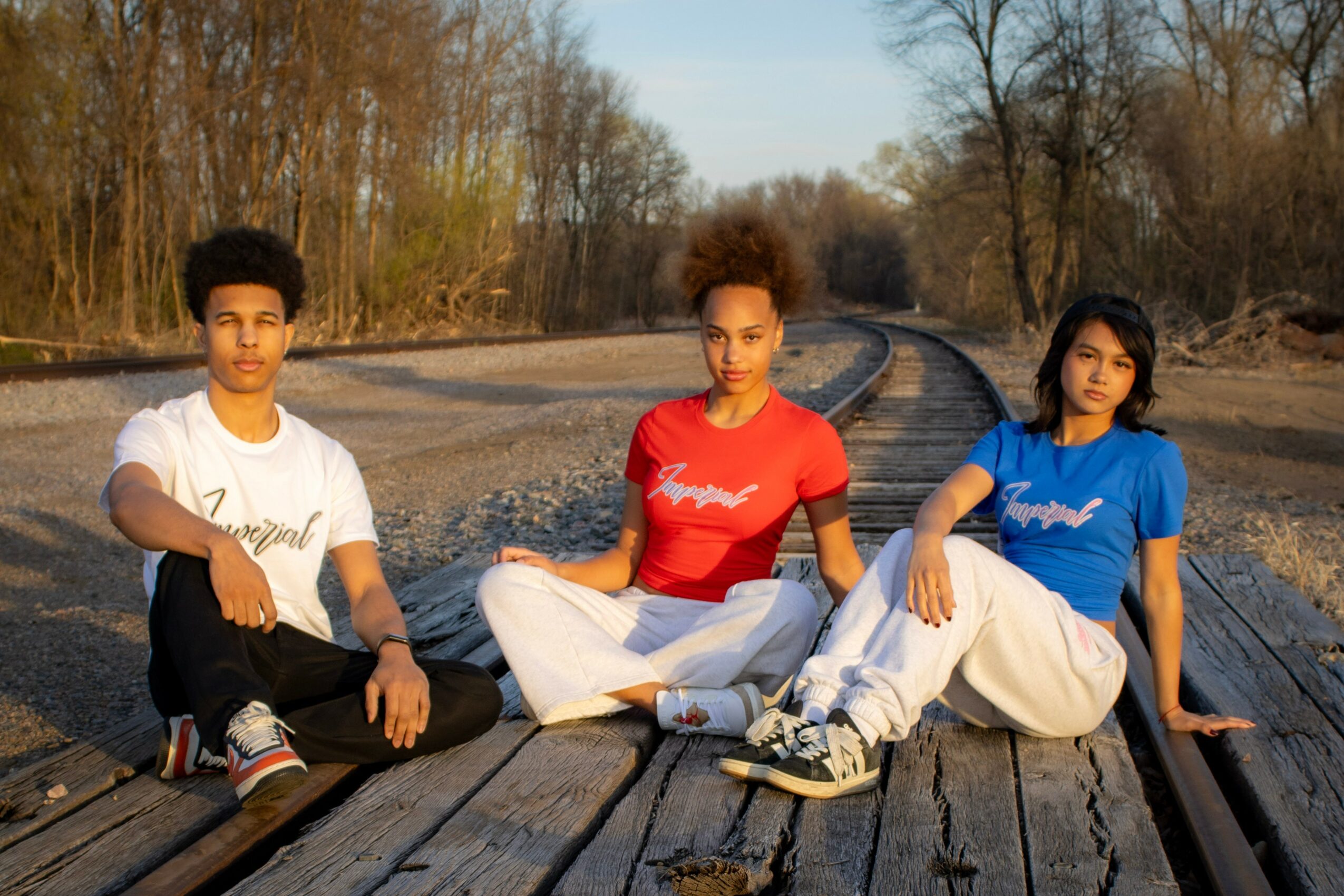 Three young people sitting on a wooden railway track outdoors, with trees and sky in the background.