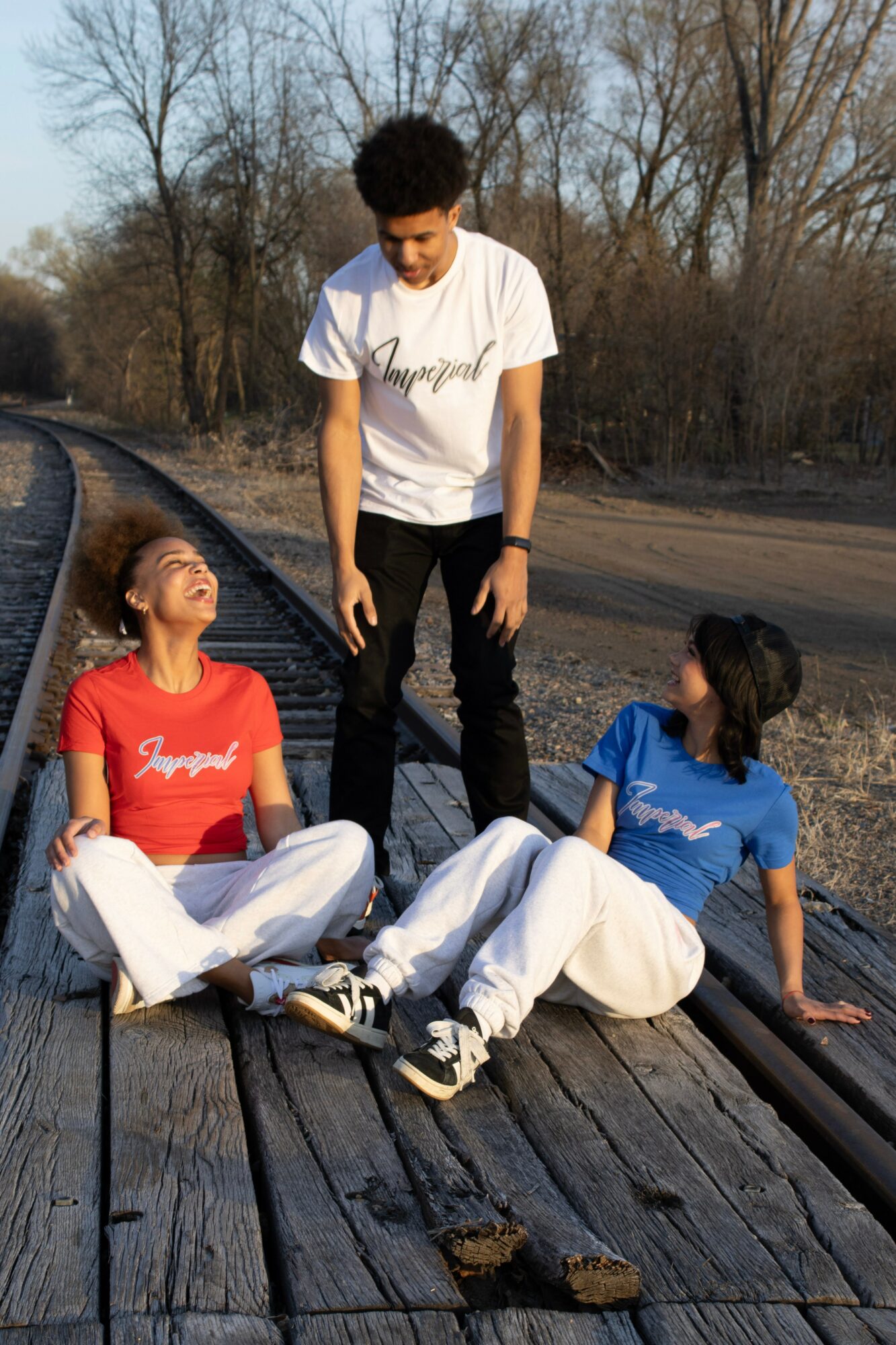 Three young people on a wooden railroad track, two sitting and one standing, outdoors with trees in background.
