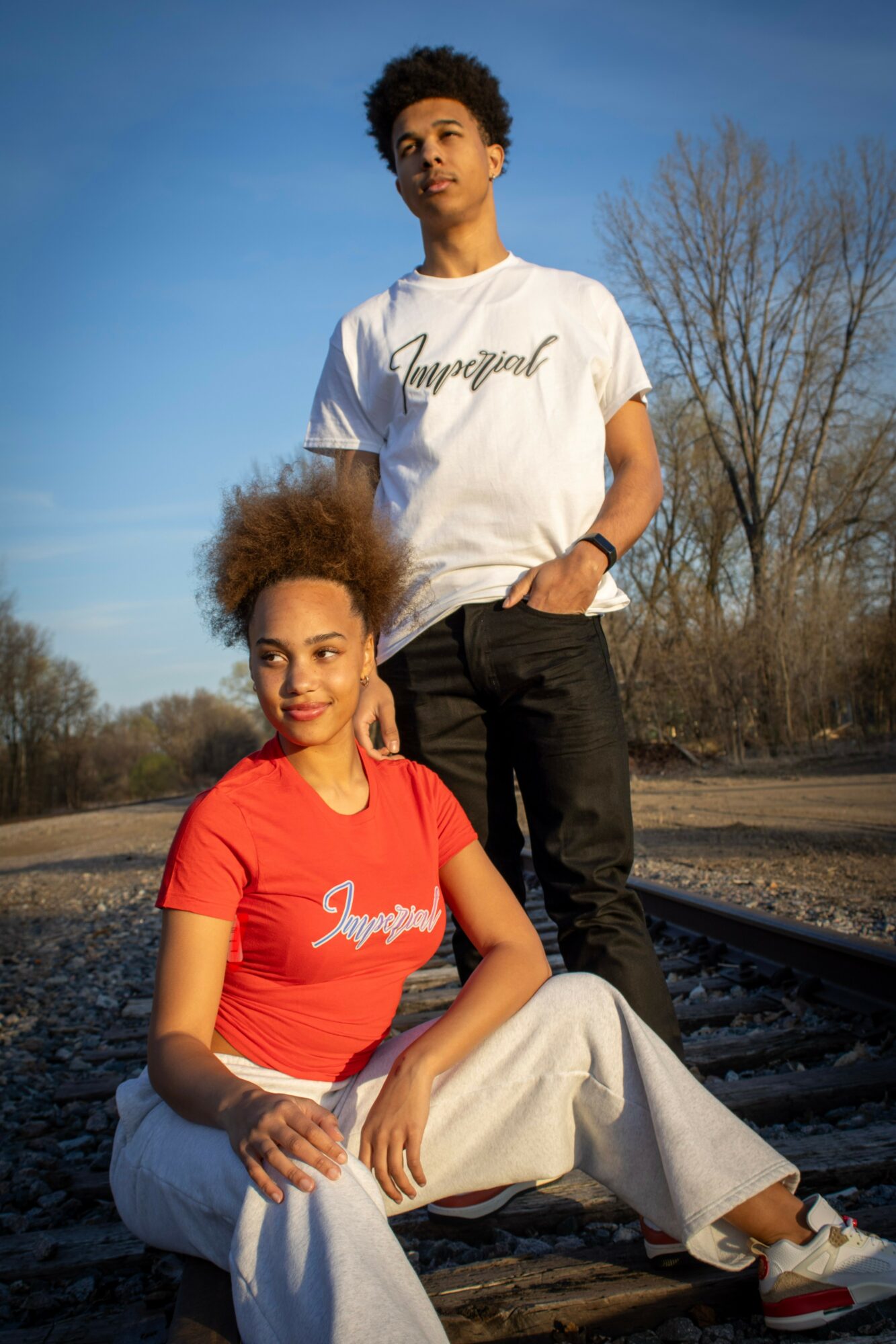 A young man and woman pose outdoors near train tracks, with trees and a blue sky in the background.