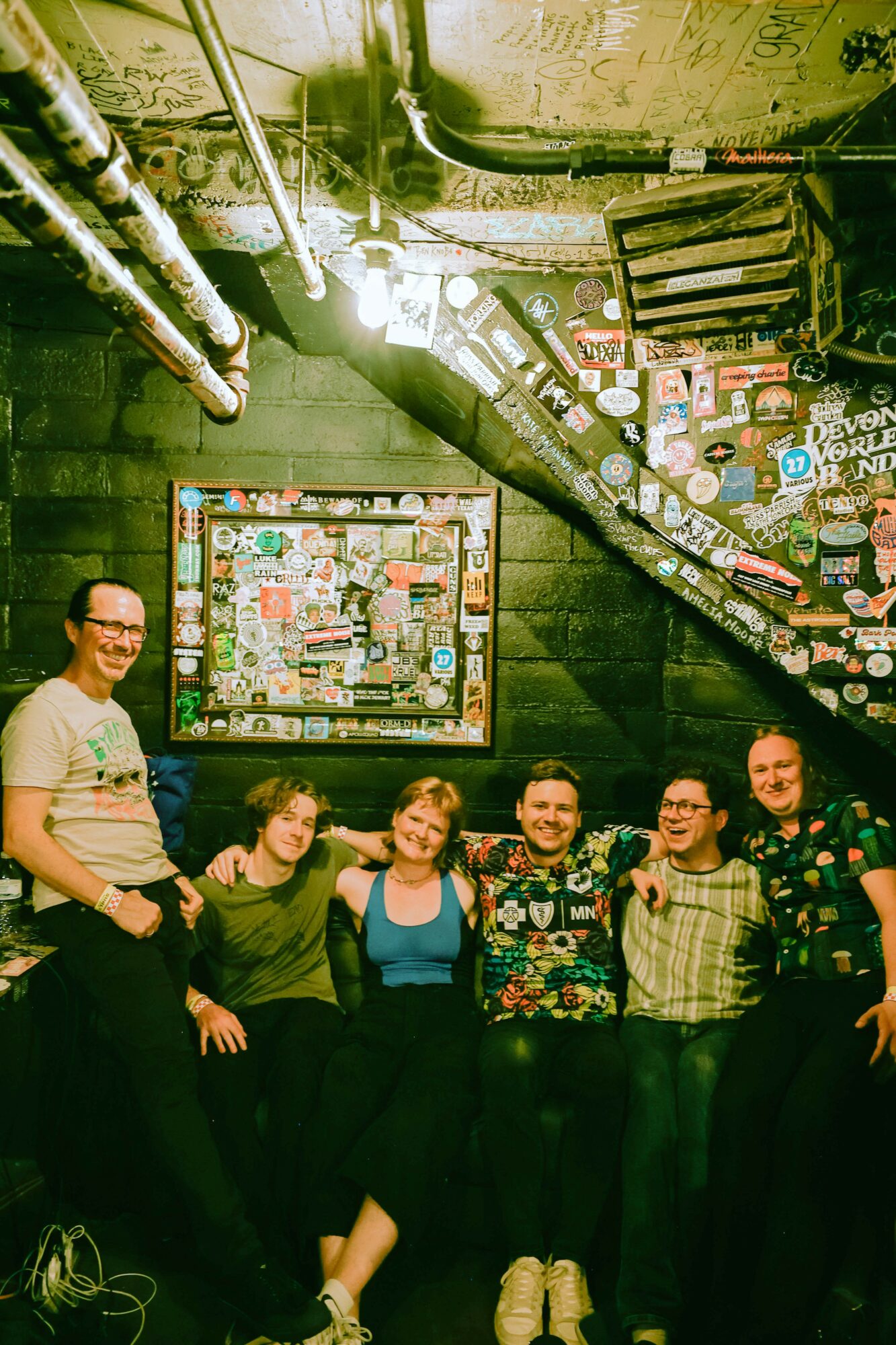 Group of six people smiling and posing in a dimly lit room with graffiti-covered walls and ceiling, some wearing glasses.
