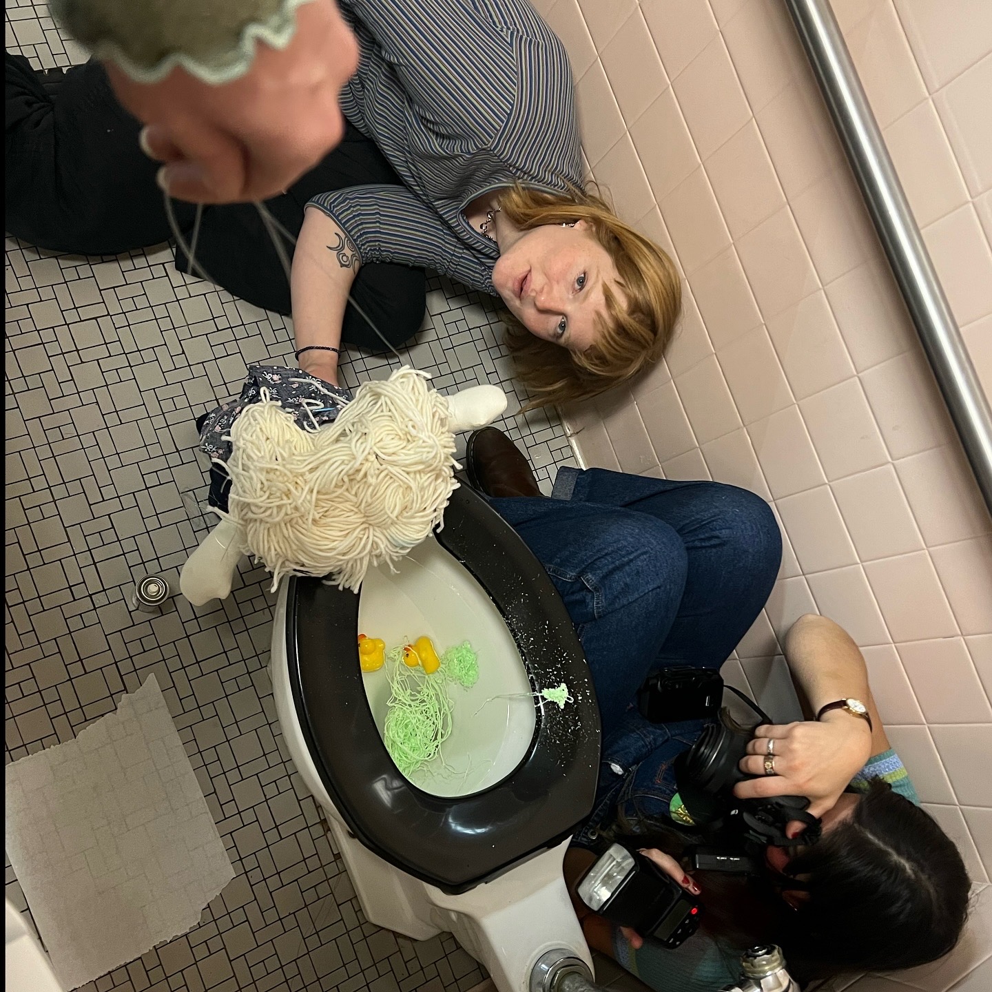 Woman lying on bathroom floor with a dog, woman holding camera, tiled wall background, bathroom setting.