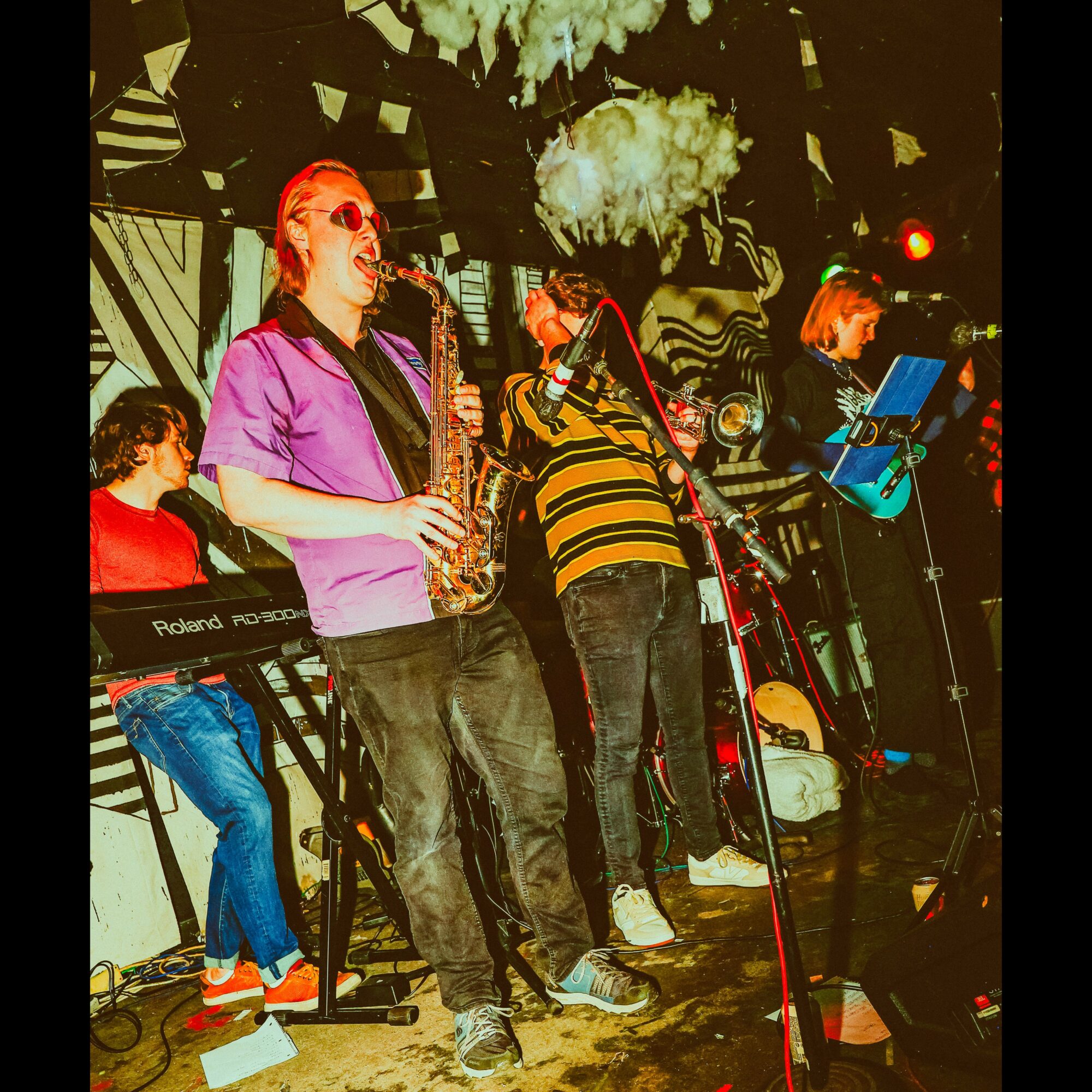 Group of musicians performing on stage with instruments, colorful lighting, and a decorated background.