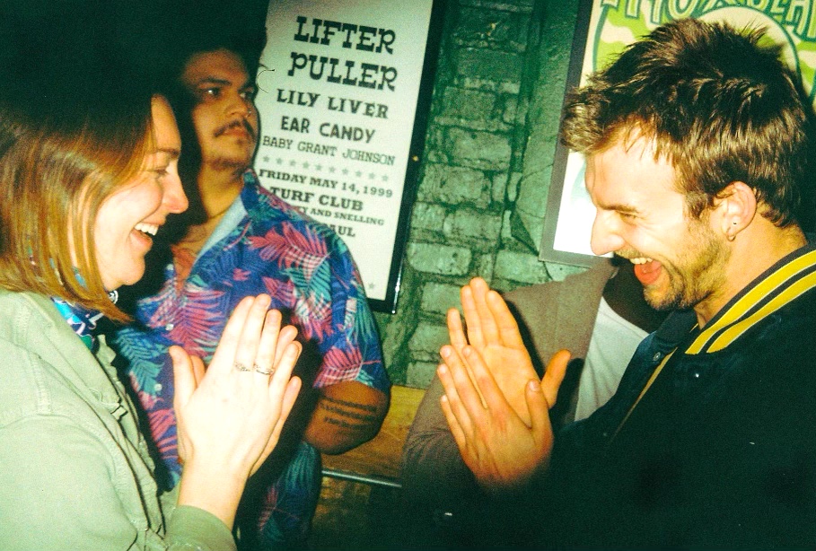 Two people smiling and greeting each other with hands pressed together, in a lively indoor setting.