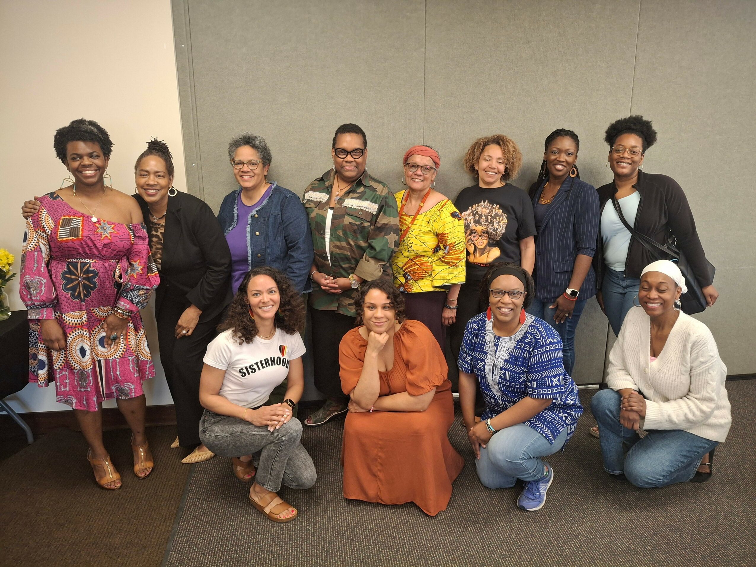 Group of twelve diverse women standing and sitting against a plain wall, smiling and posing for the photo.