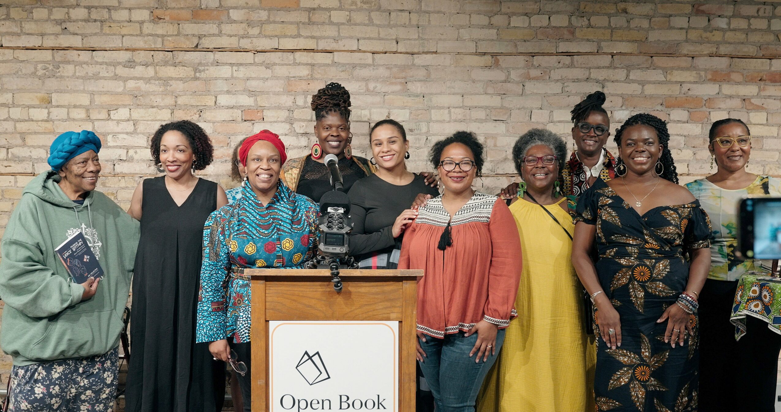 Group of women standing behind a podium with a sign that says 'Open Book' in front of a brick wall.