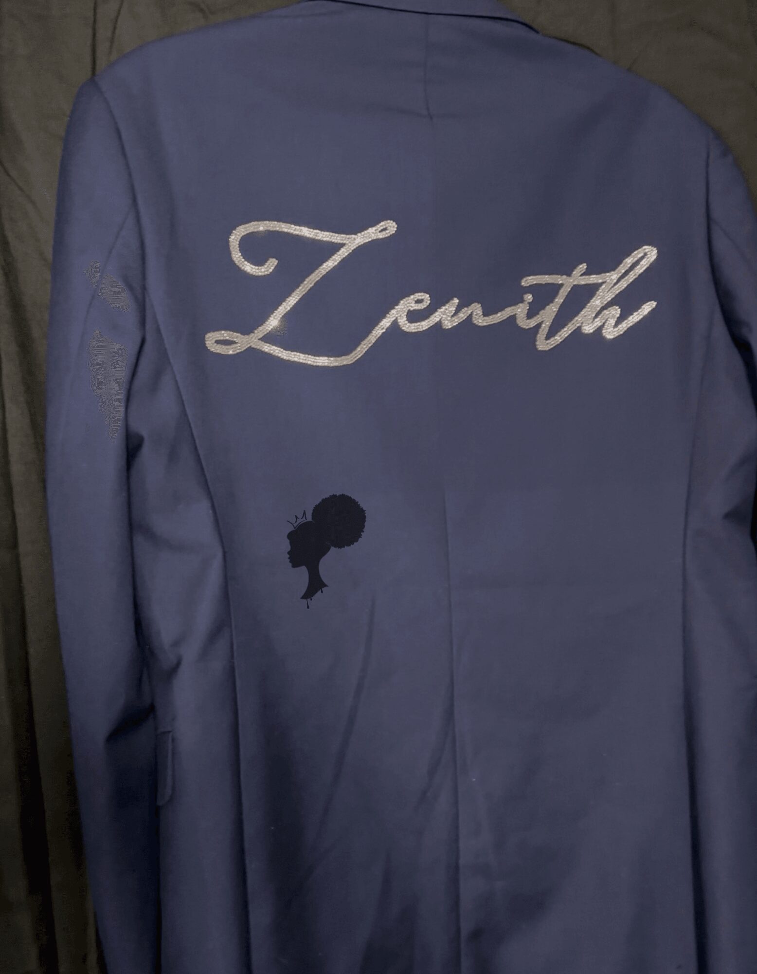 Back of a blue jacket with the word 'Zenith' embroidered in cursive and a small black silhouette of a person's head.