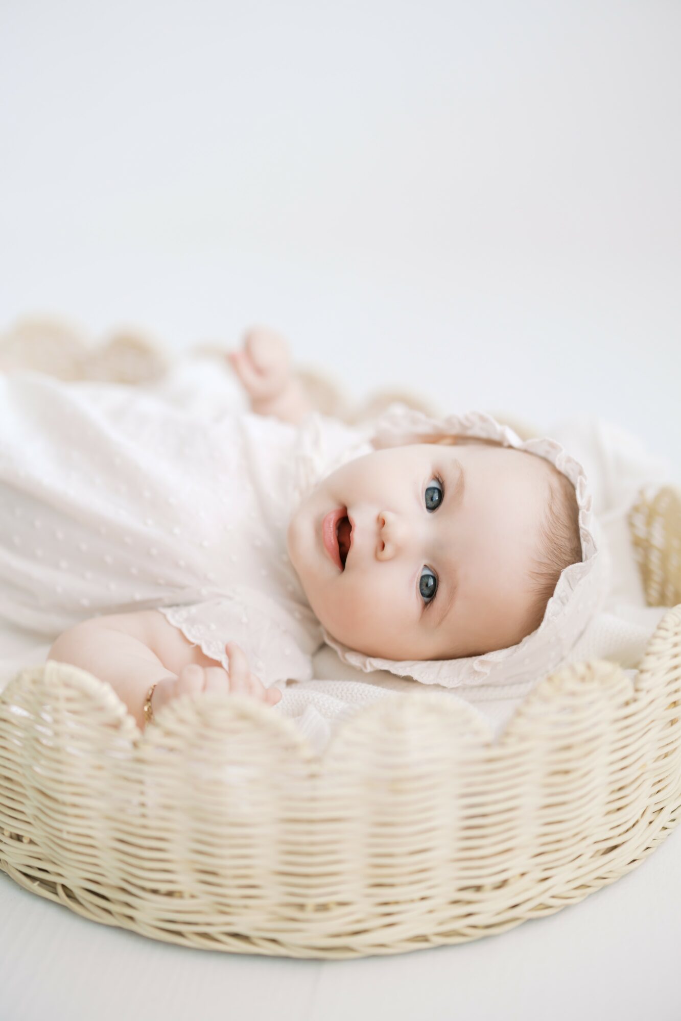 Baby lying in a woven basket, looking up with wide eyes, wearing a light-colored outfit, on a white background.