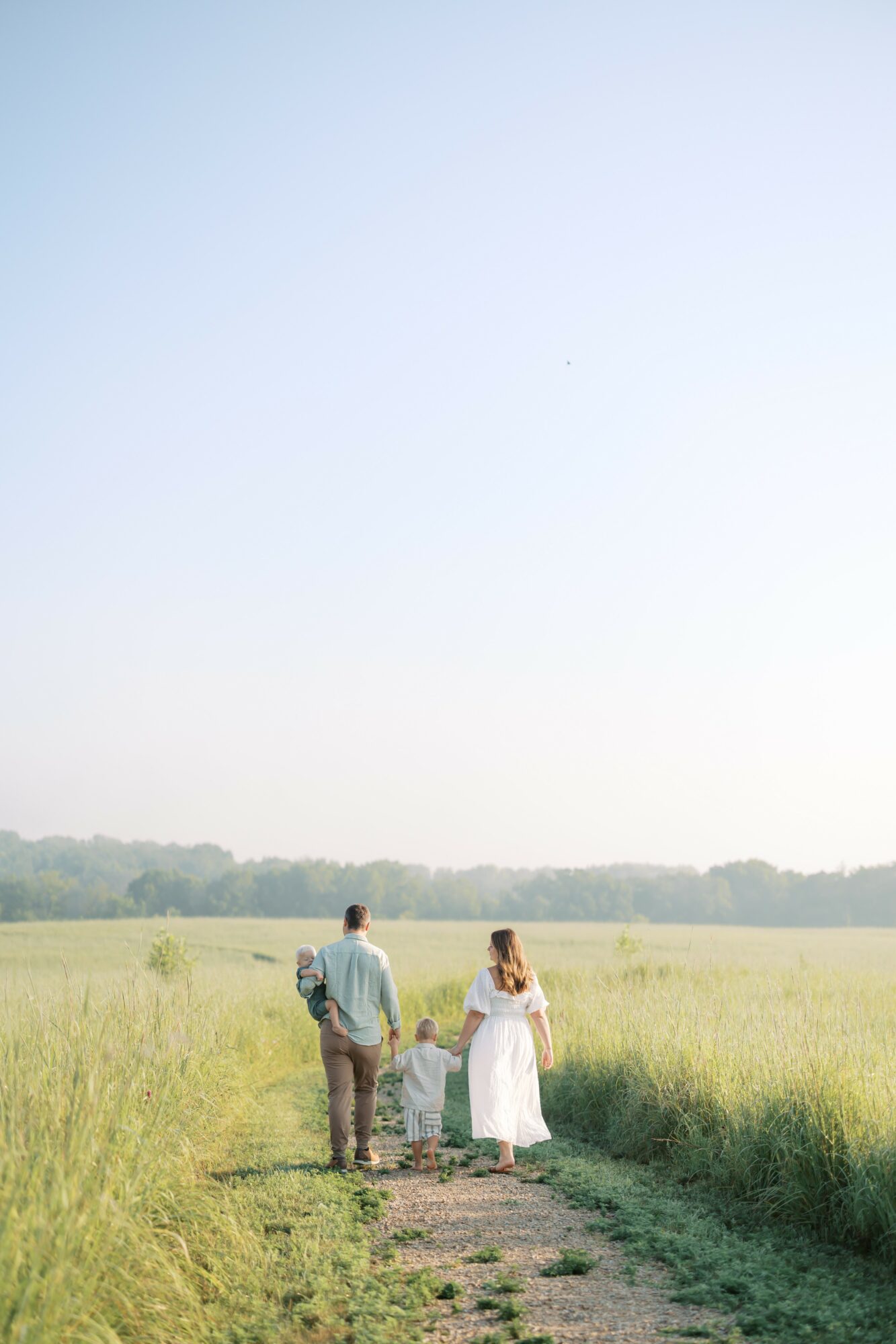 A man and woman walk along a grassy path in a field under a clear sky, holding hands and facing away from the camera.