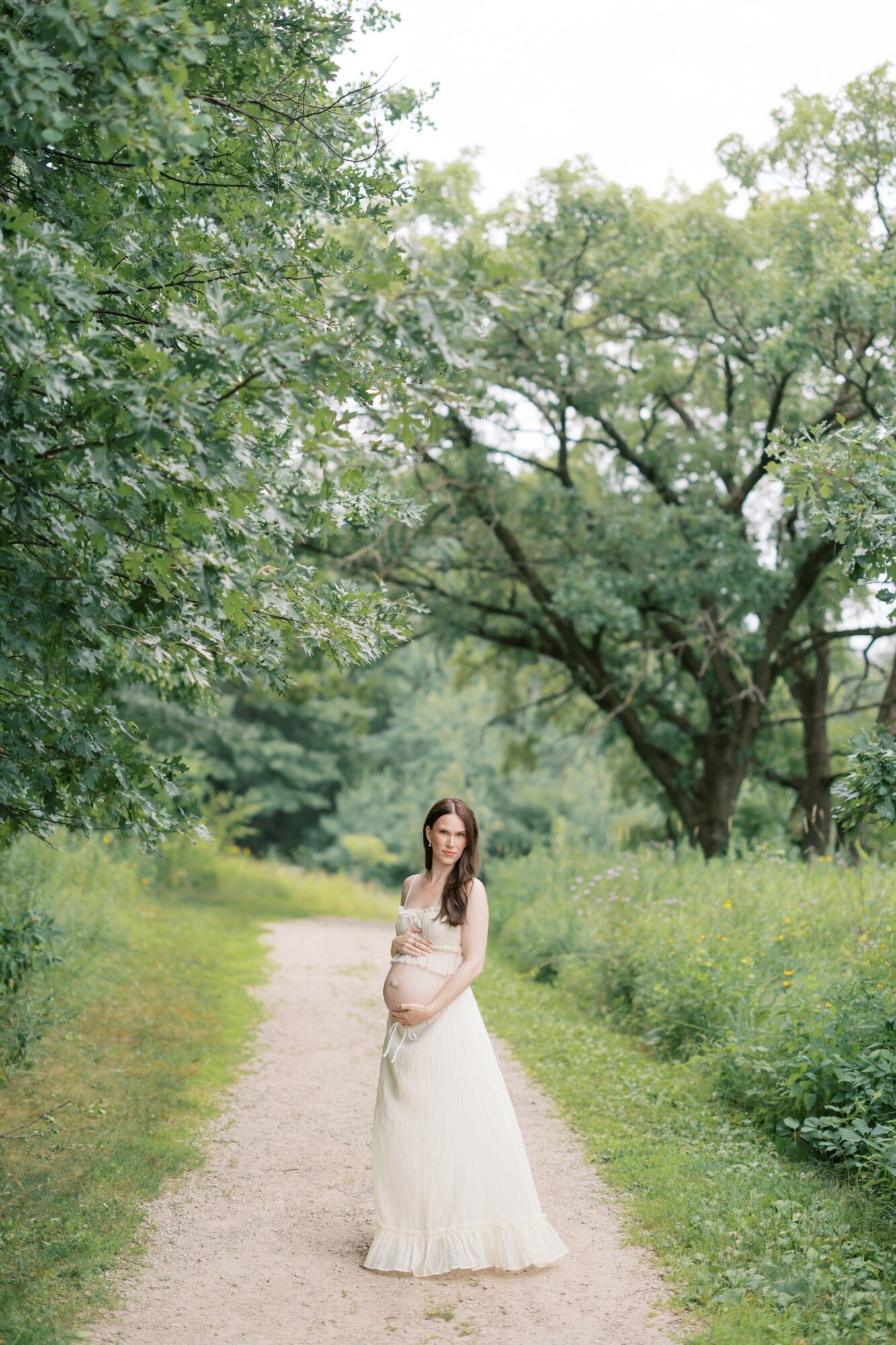 Woman in a white dress standing on a dirt path surrounded by green trees and grass, overcast sky