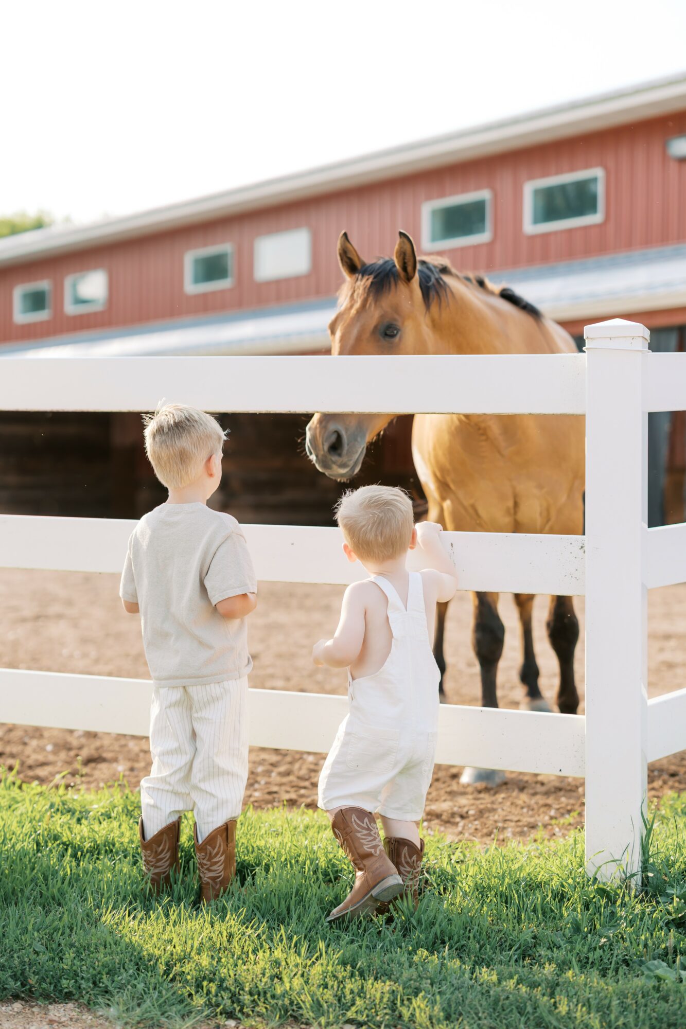 Two children in white outfits and boots stand near a white fence, observing a horse in a stable yard.