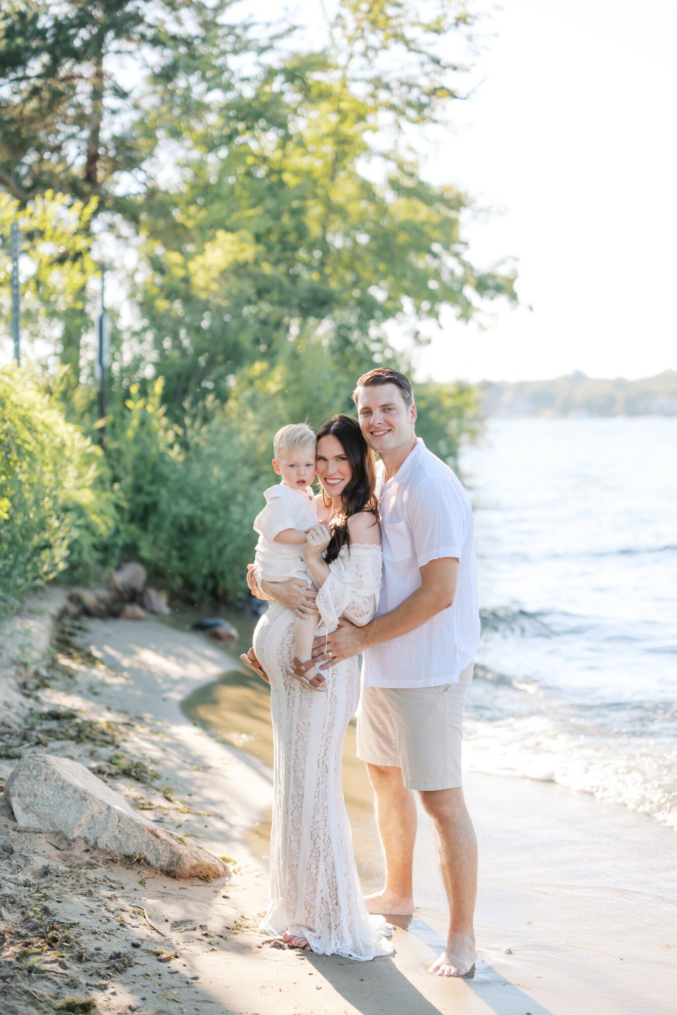 Family of three standing on a beach near water, trees in background, sunny day, smiling.