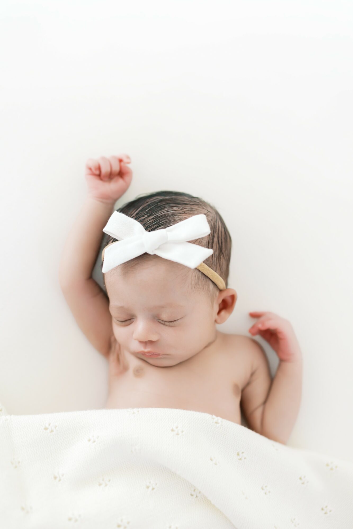 Baby with a white bow headband sleeping, one arm raised, in a white bathtub or bed, neutral background.