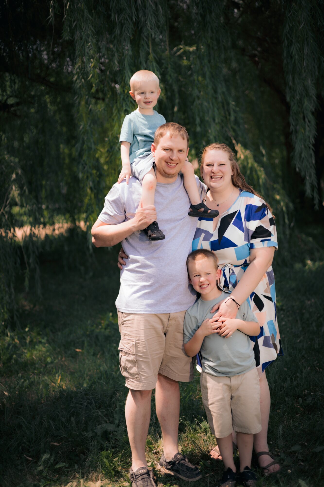 Family of four outdoors, one child on father's shoulders, trees in background, smiling faces, casual clothing.