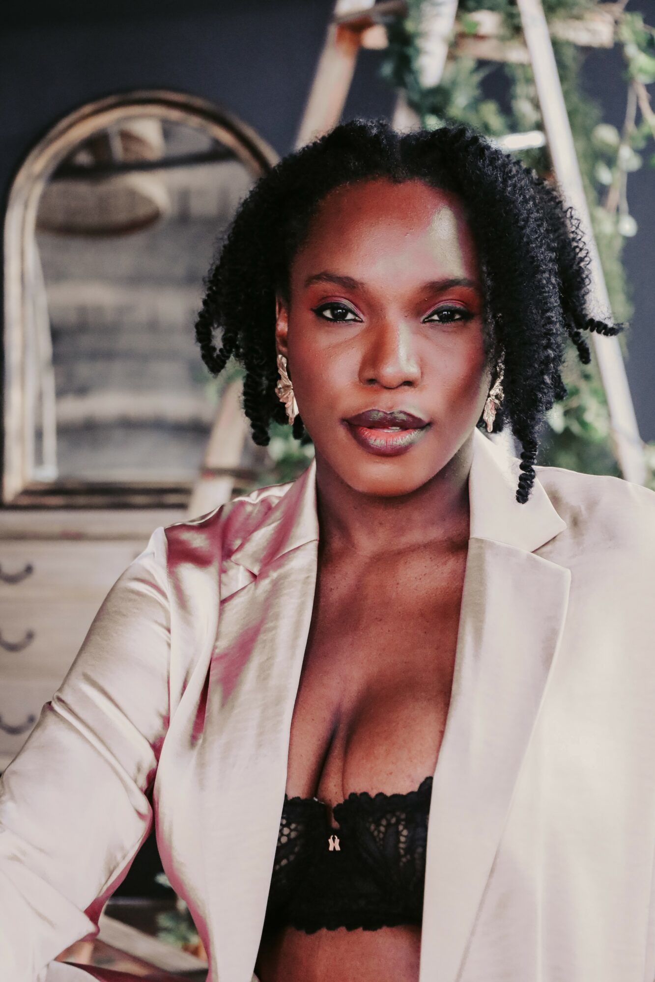 Woman with natural hair wearing a satin blazer and black lace top, sitting indoors with a mirror and plant in background.