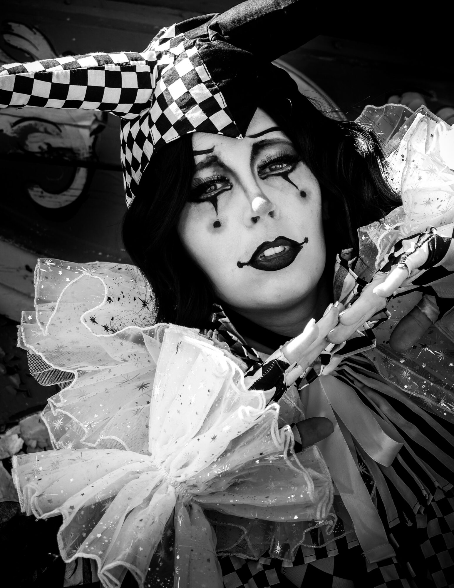 Person dressed as a clown with makeup, checkered hat, and ruffled costume, lying down with a dark background.