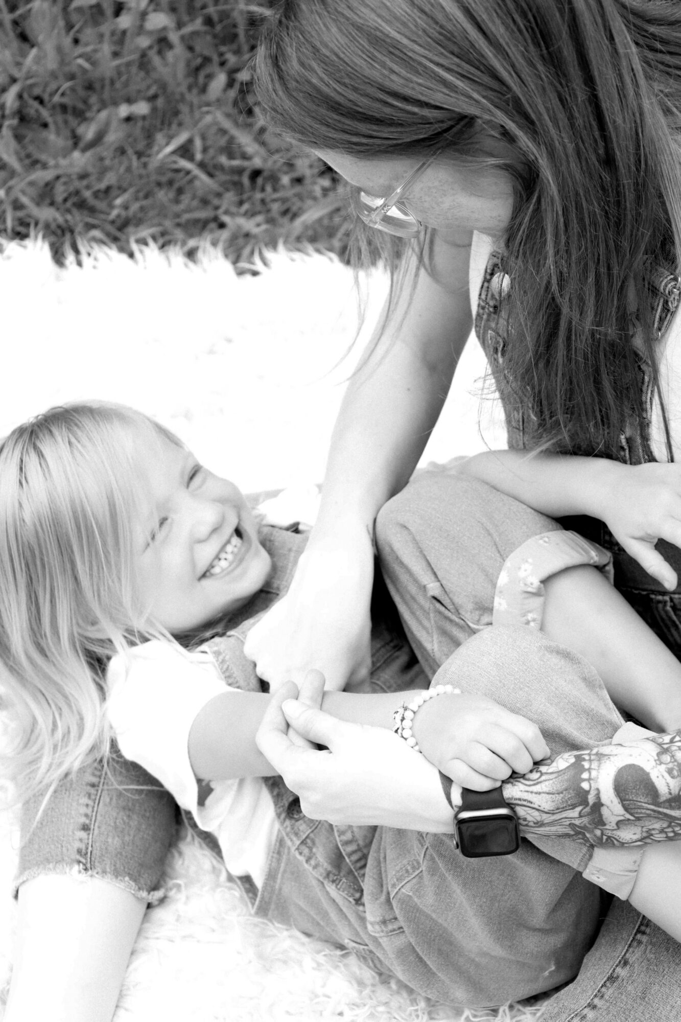Woman and girl lying on grass, smiling and holding hands, outdoor setting, black and white photo.