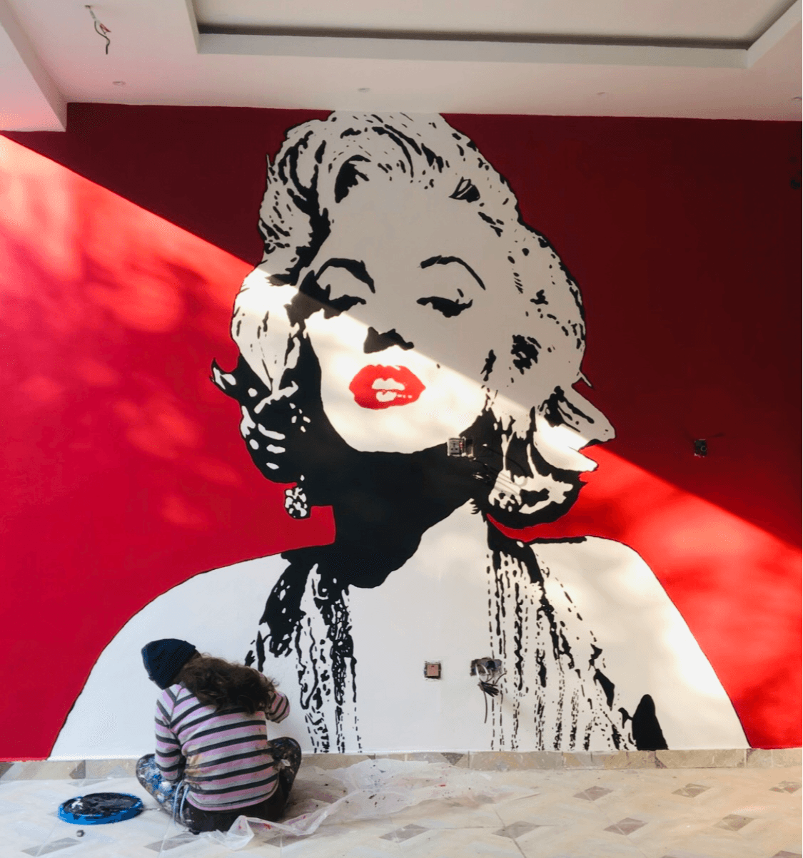 Person sitting on the floor in front of a large Marilyn Monroe mural on a red wall.