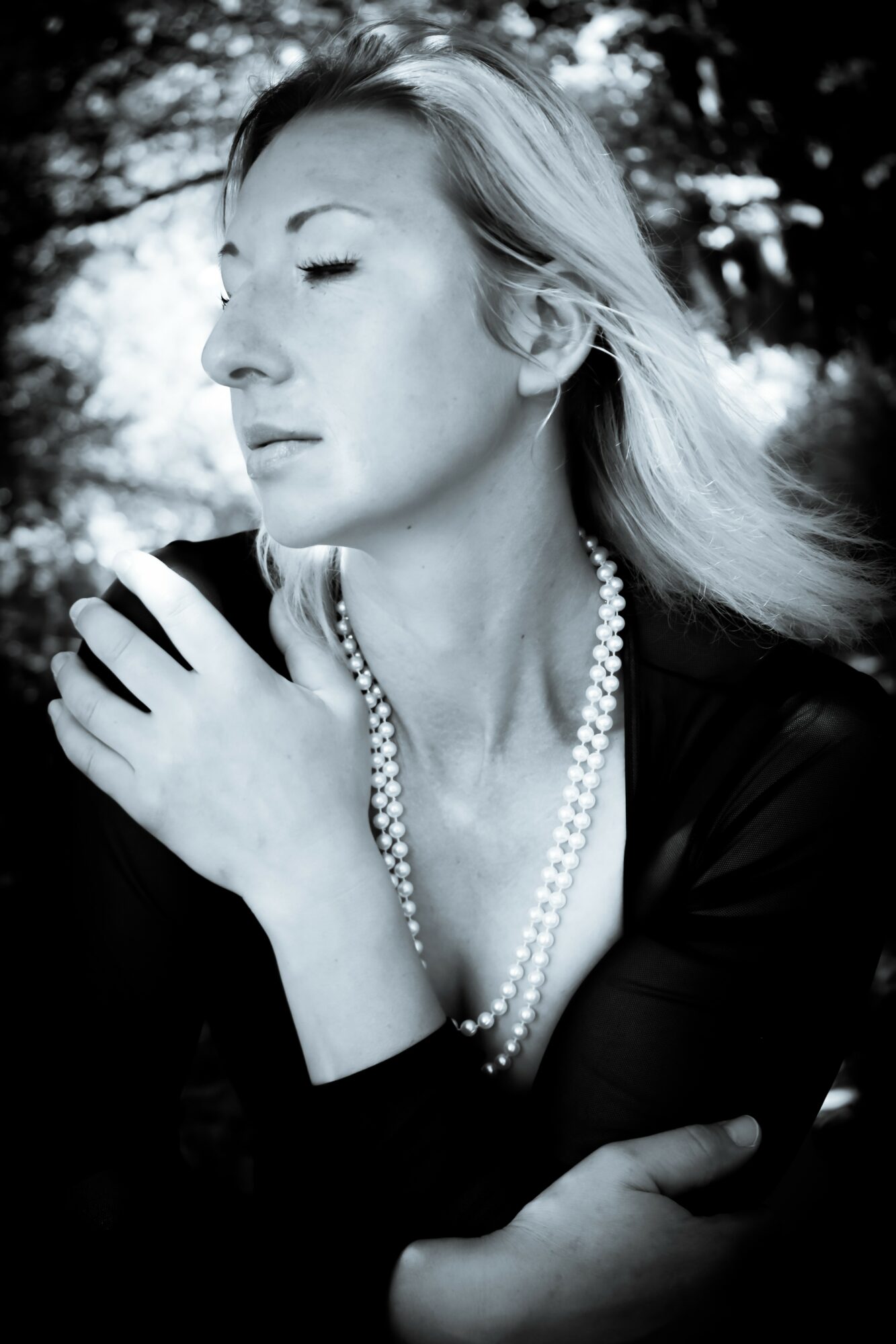 Woman with closed eyes and pearl necklace outdoors, black and white photo, serene expression, hand on shoulder.