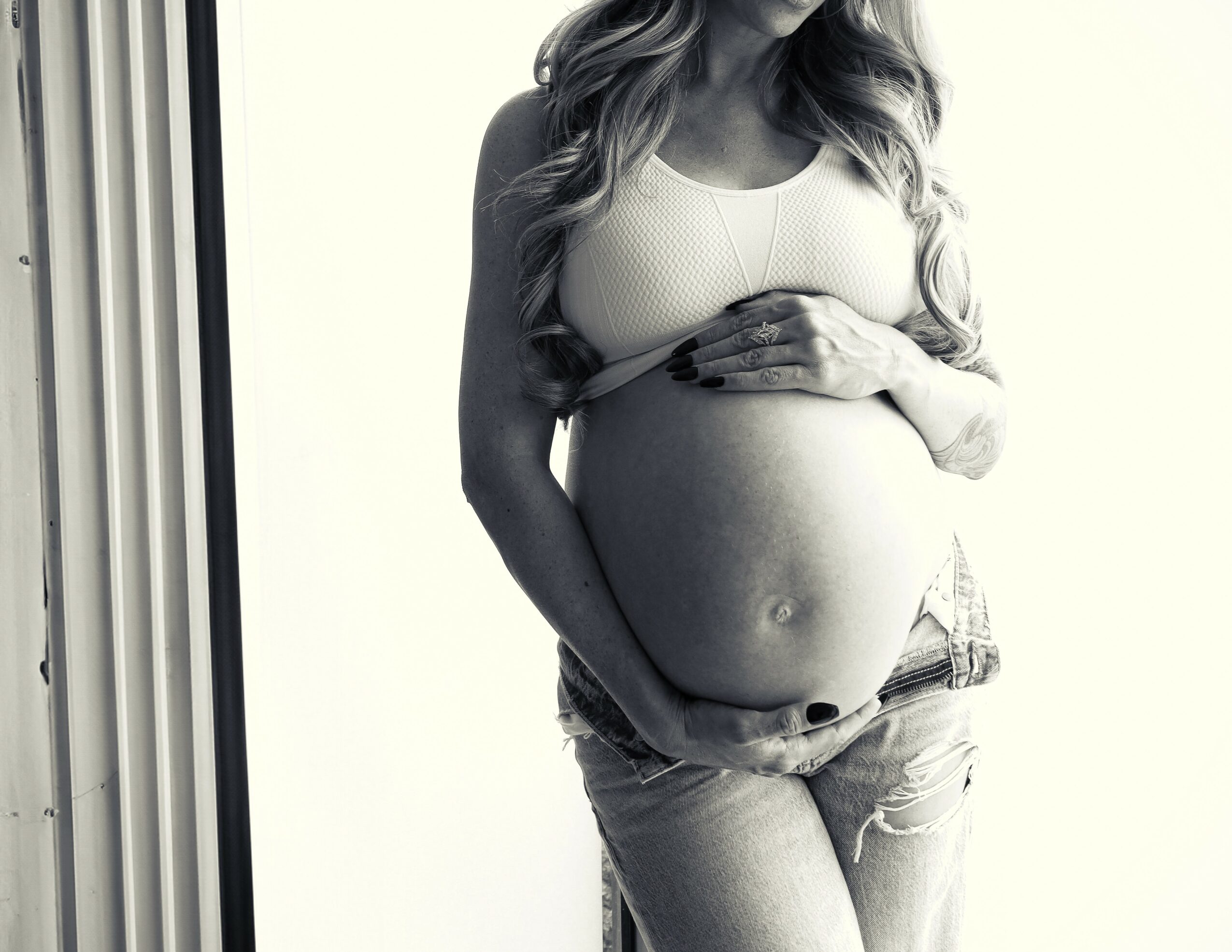 Pregnant woman with long hair, wearing a sleeveless top and jeans, holding her belly with both hands.