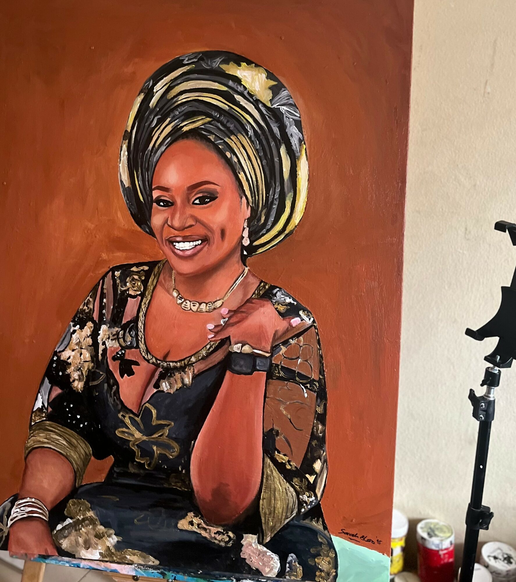 Woman wearing traditional African attire and headwrap, smiling, sitting with her hand on her chin, against a brown background.