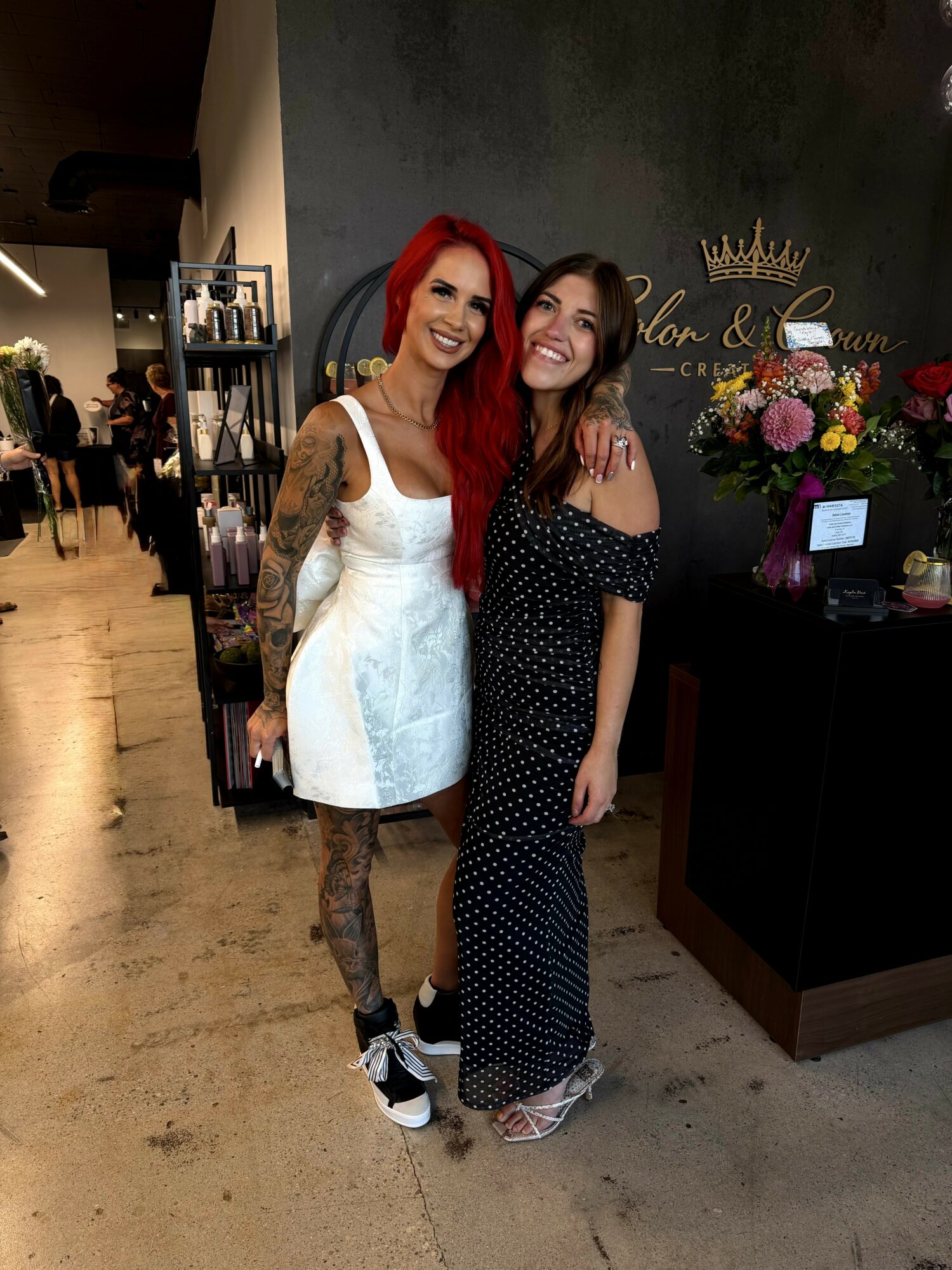 Two women smiling indoors, one with red hair and tattoos, the other with dark hair in a black dress, standing near a floral display.