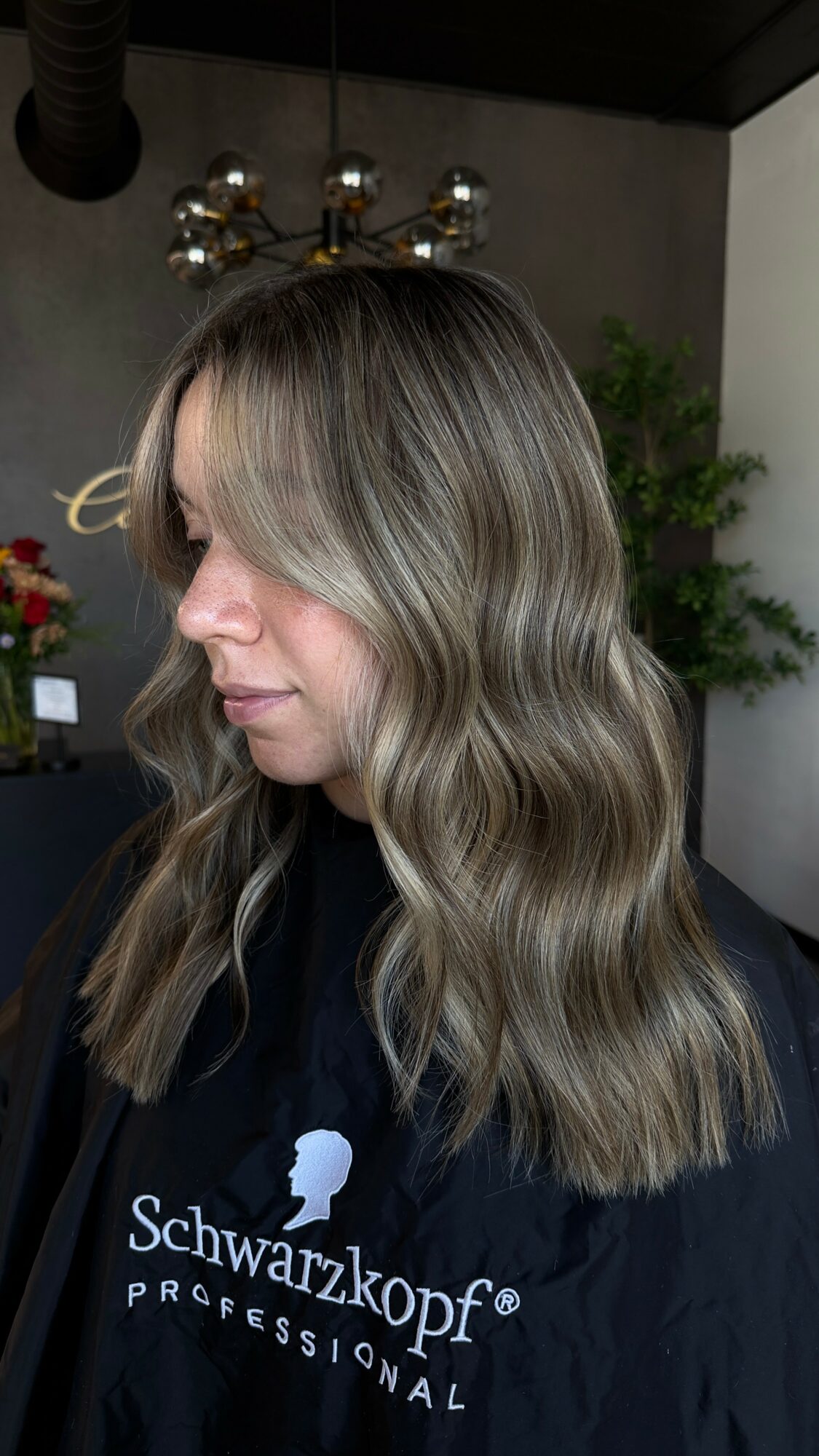 Woman with wavy, shoulder-length hair in a salon, with a chandelier and plant in the background.