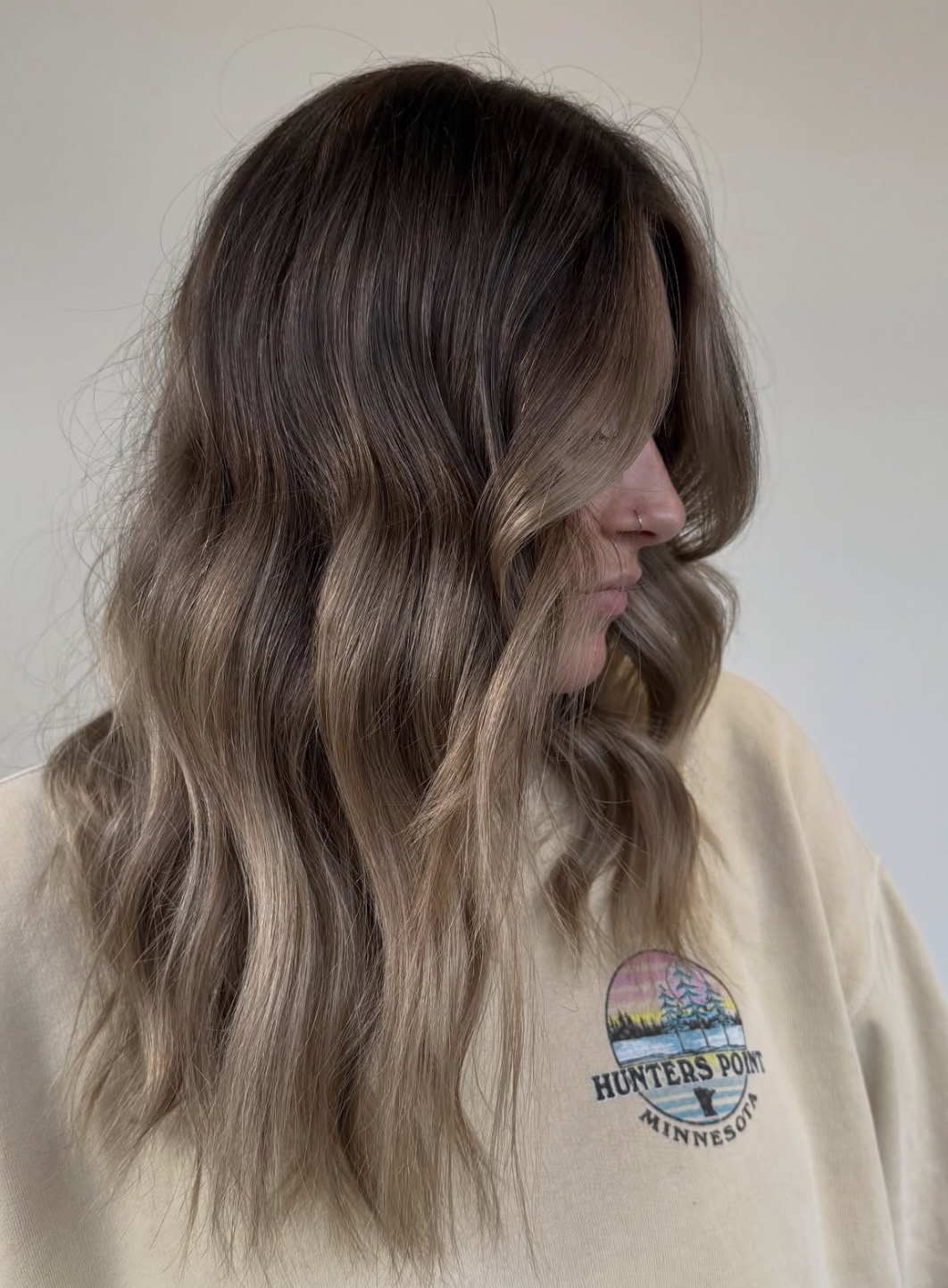Side view of a woman with long, wavy hair, wearing a beige shirt with a logo, against a plain background.