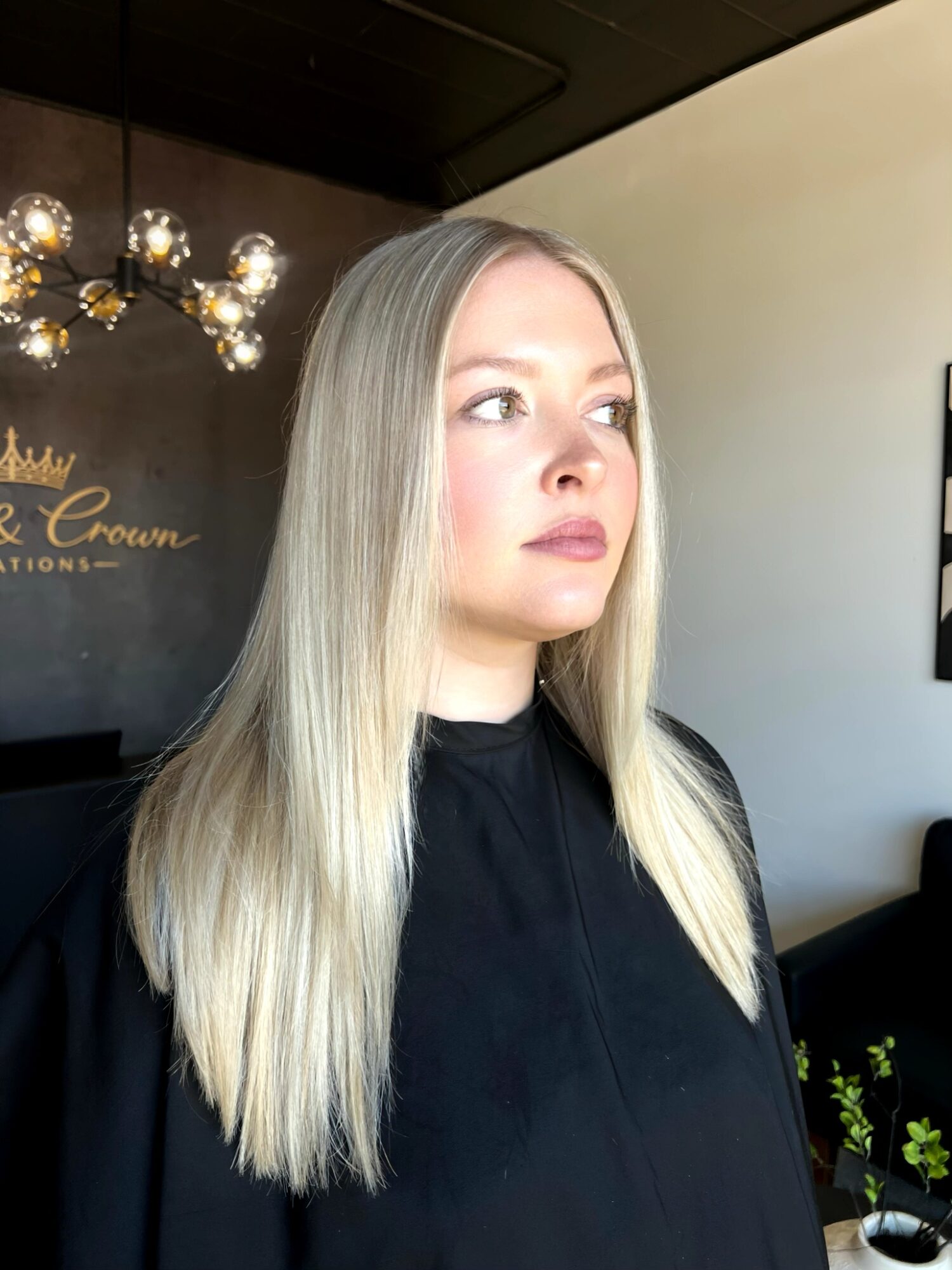 Young woman with long blonde hair and light makeup, wearing a black top, looking to the side indoors.