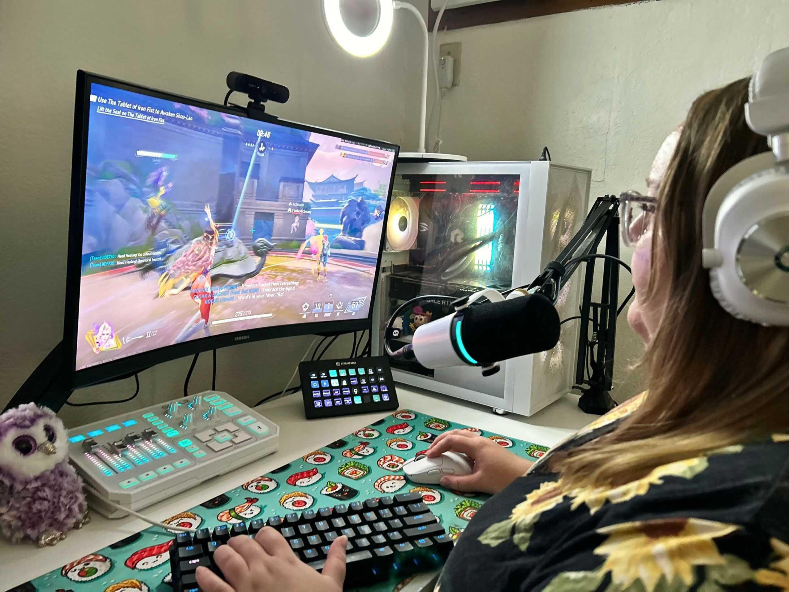 Person wearing headphones playing a video game on a computer with a large monitor, keyboard, and microphone on desk.