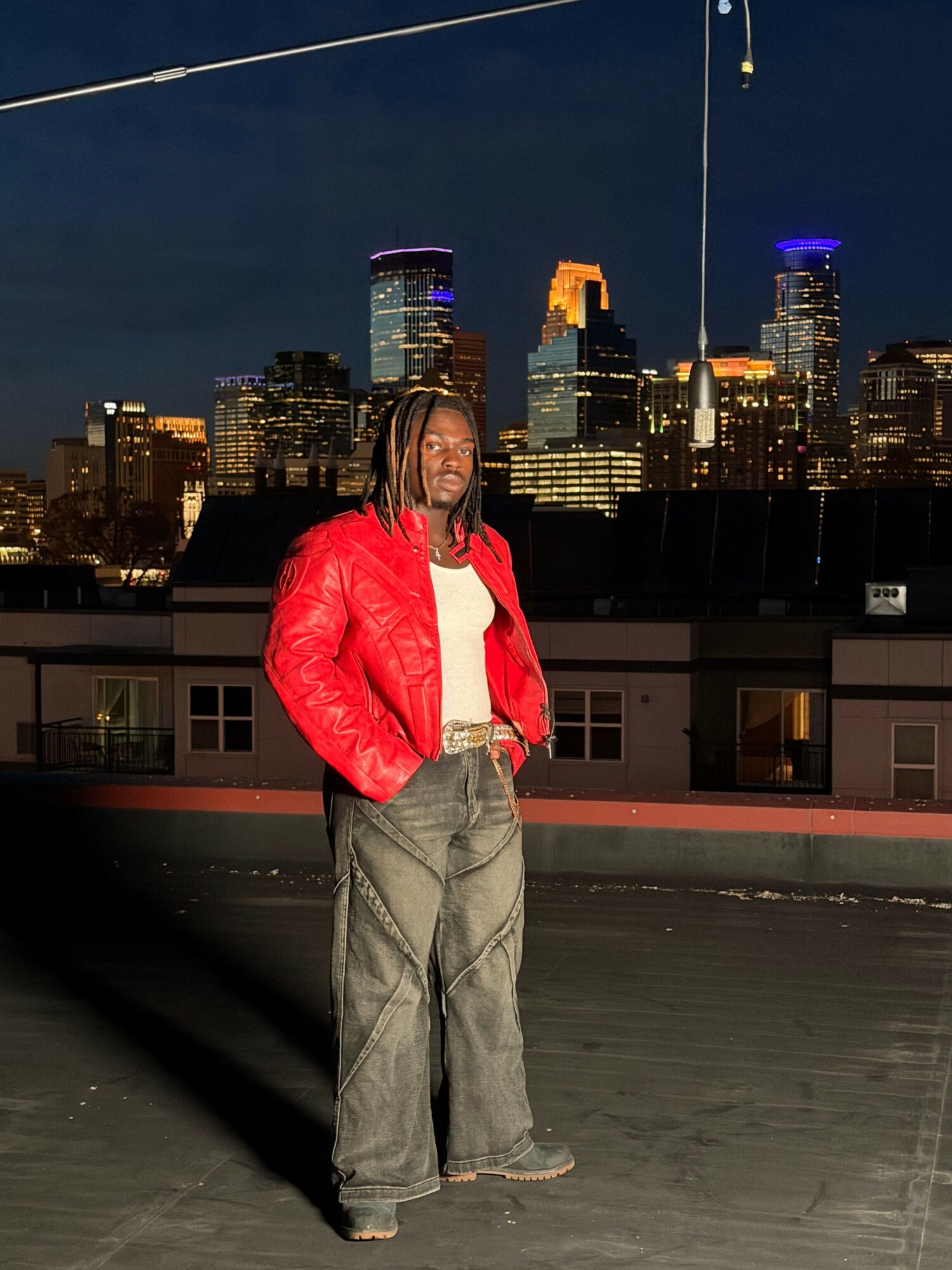 Person standing outdoors at night with city skyline in background, wearing a red jacket and gray pants.