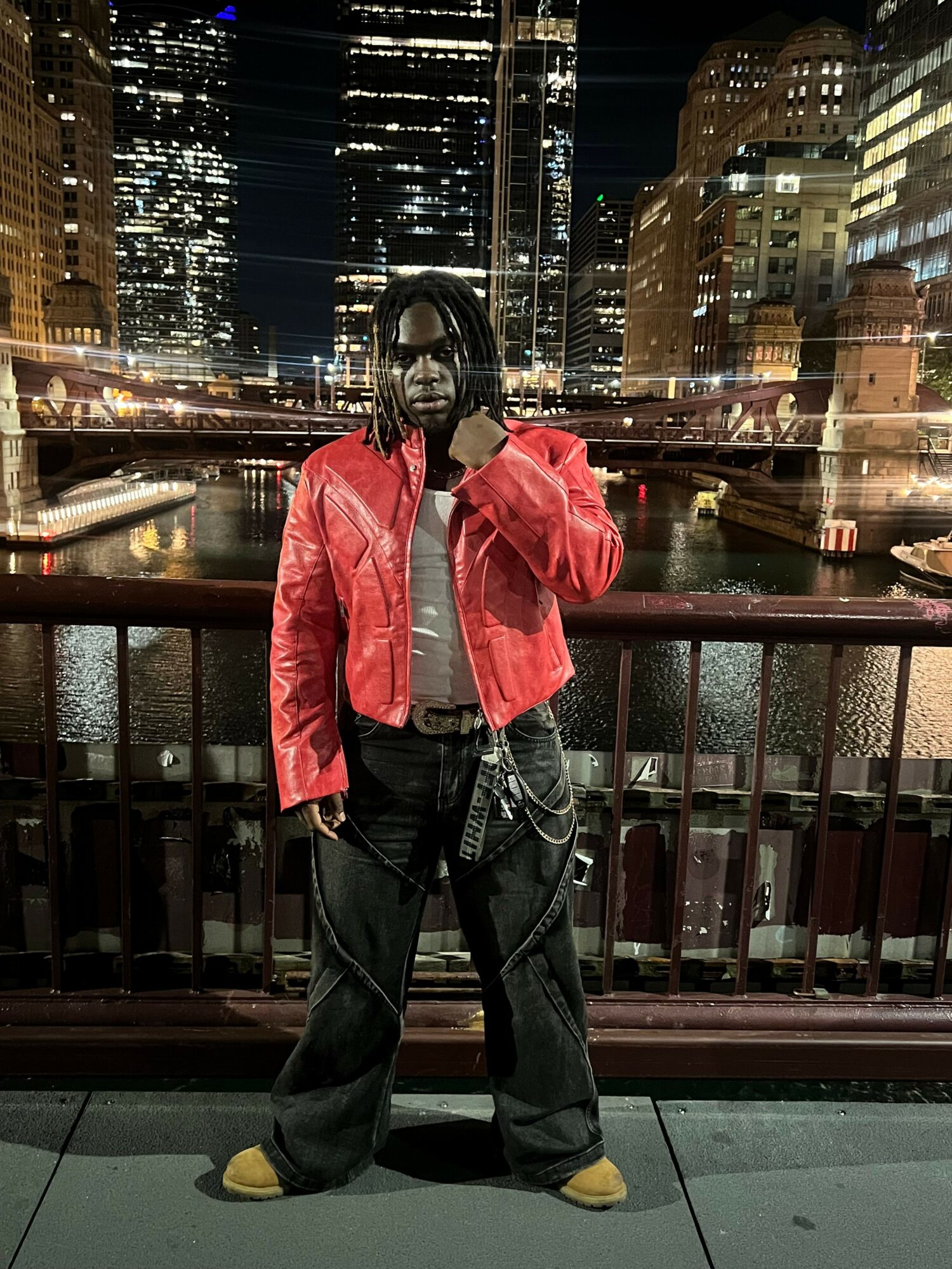 Person standing on a city sidewalk at night, wearing a red jacket, black pants, and yellow shoes, with city lights in background.