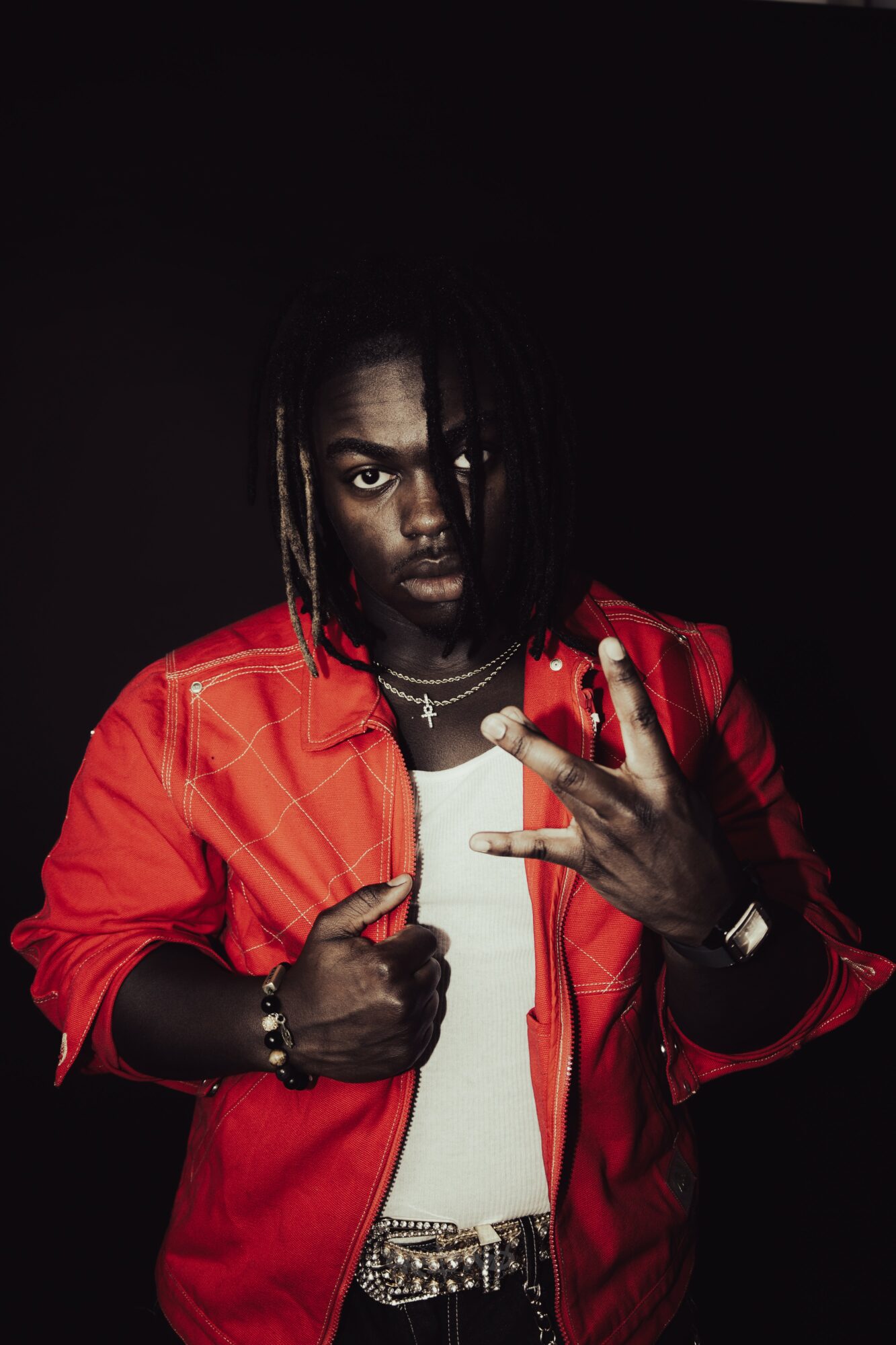 Person with dreadlocks wearing a red jacket and white shirt, making a hand gesture, against a dark background.