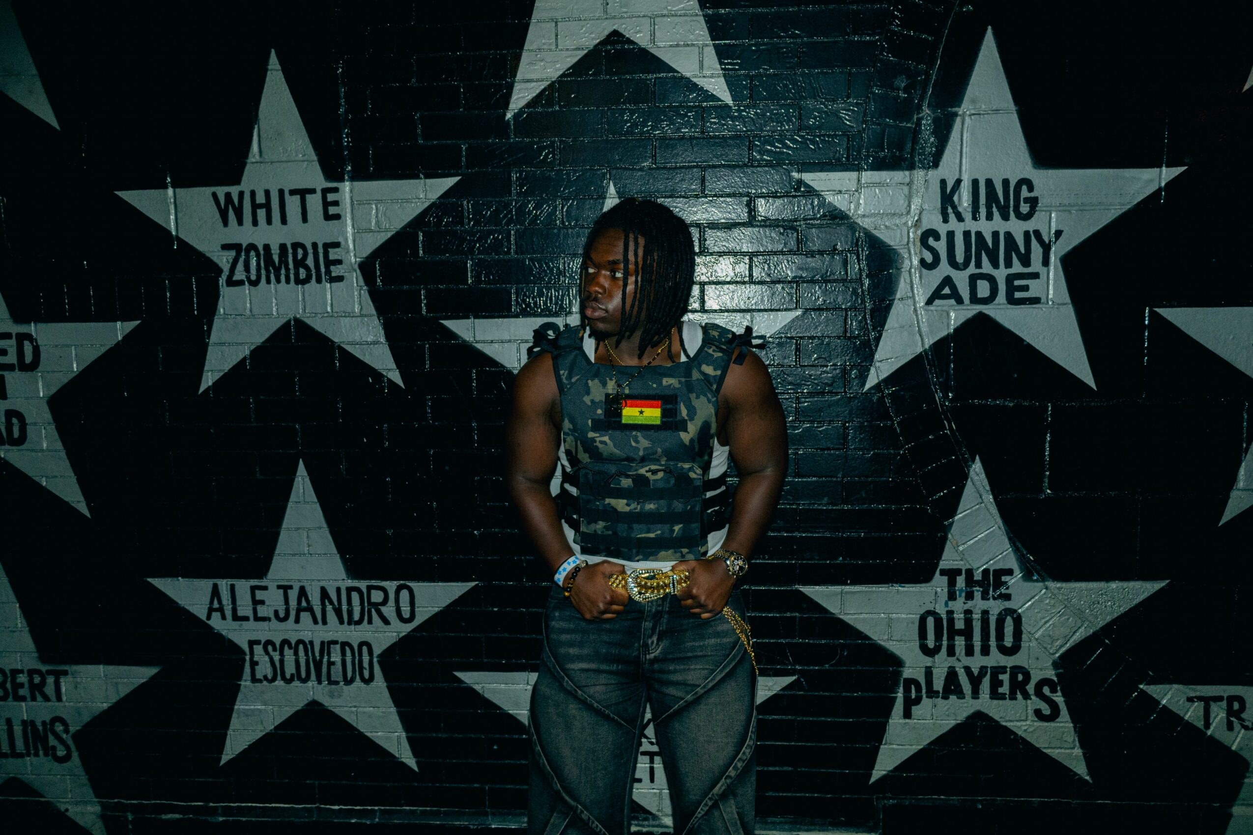 Person with dreadlocks wearing a sleeveless top and jeans stands in front of a star mural on a brick wall.