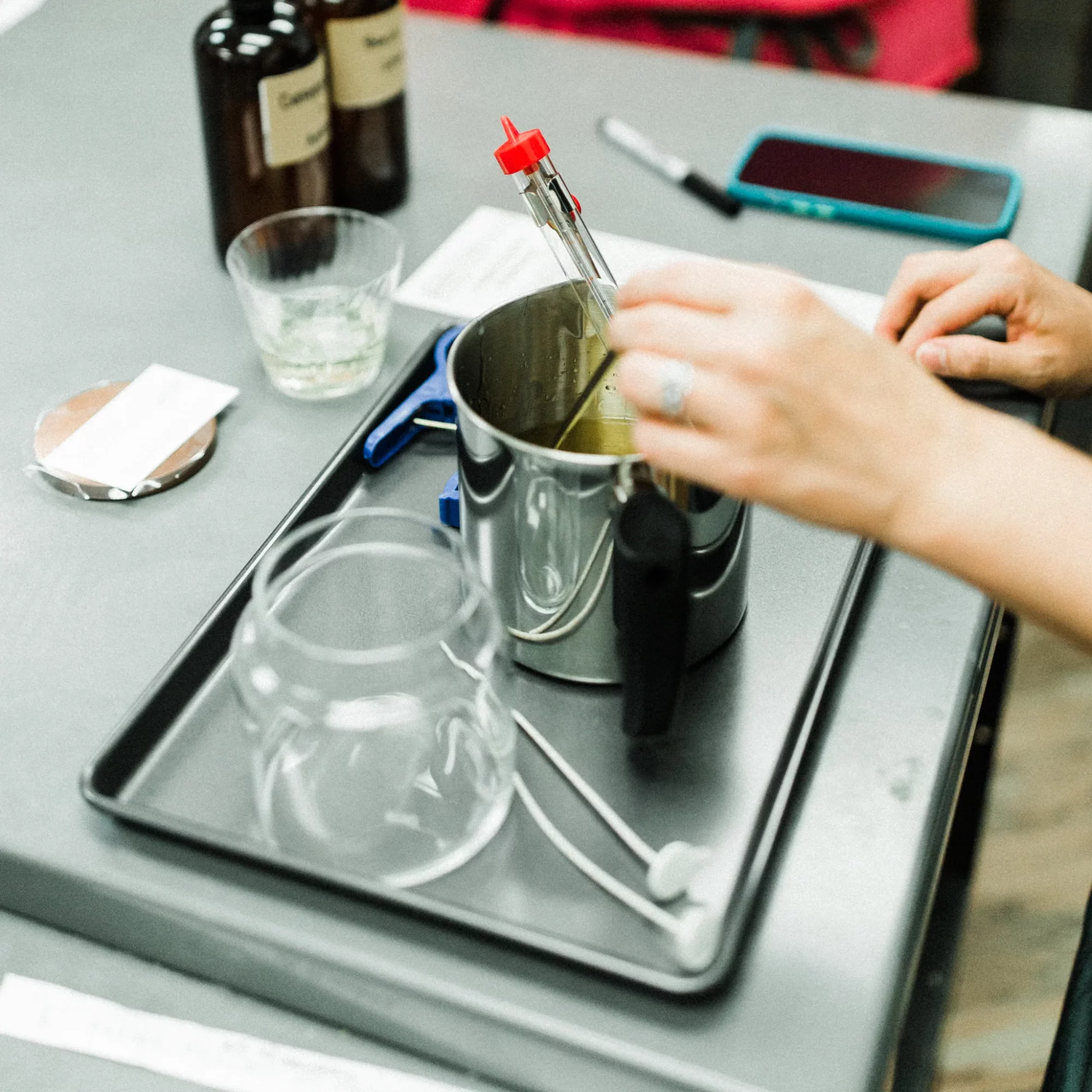 Hands holding a glass pipette over a metal container on a tray, with bottles, a glass, and a phone nearby.