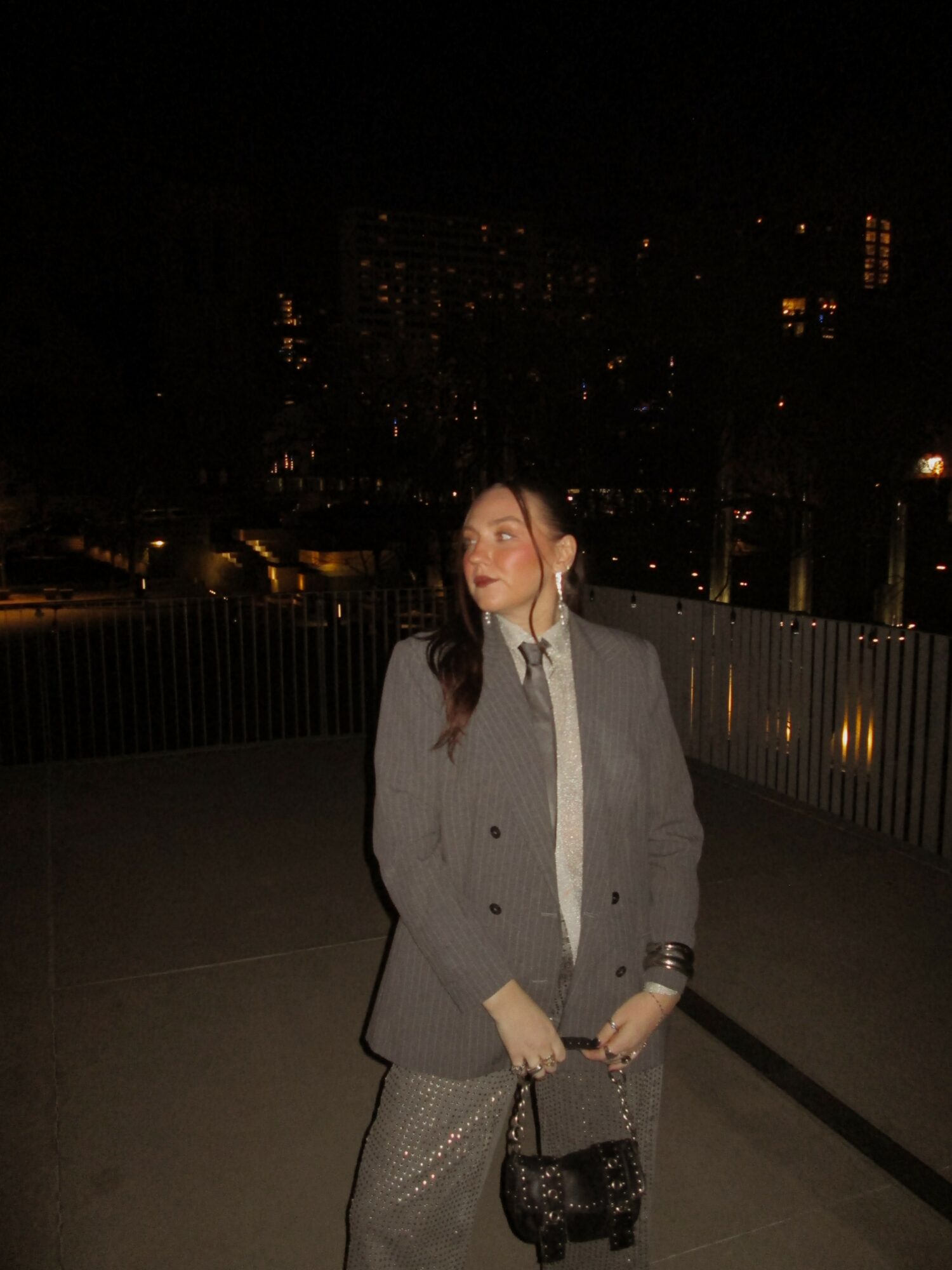 Woman in gray blazer and white shirt holding a small black bag on a balcony at night.