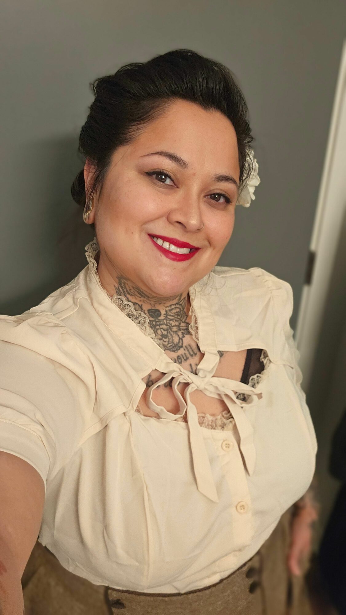 Woman with dark hair styled up, smiling, wearing a cream blouse with lace details, and a tattoo on her neck.