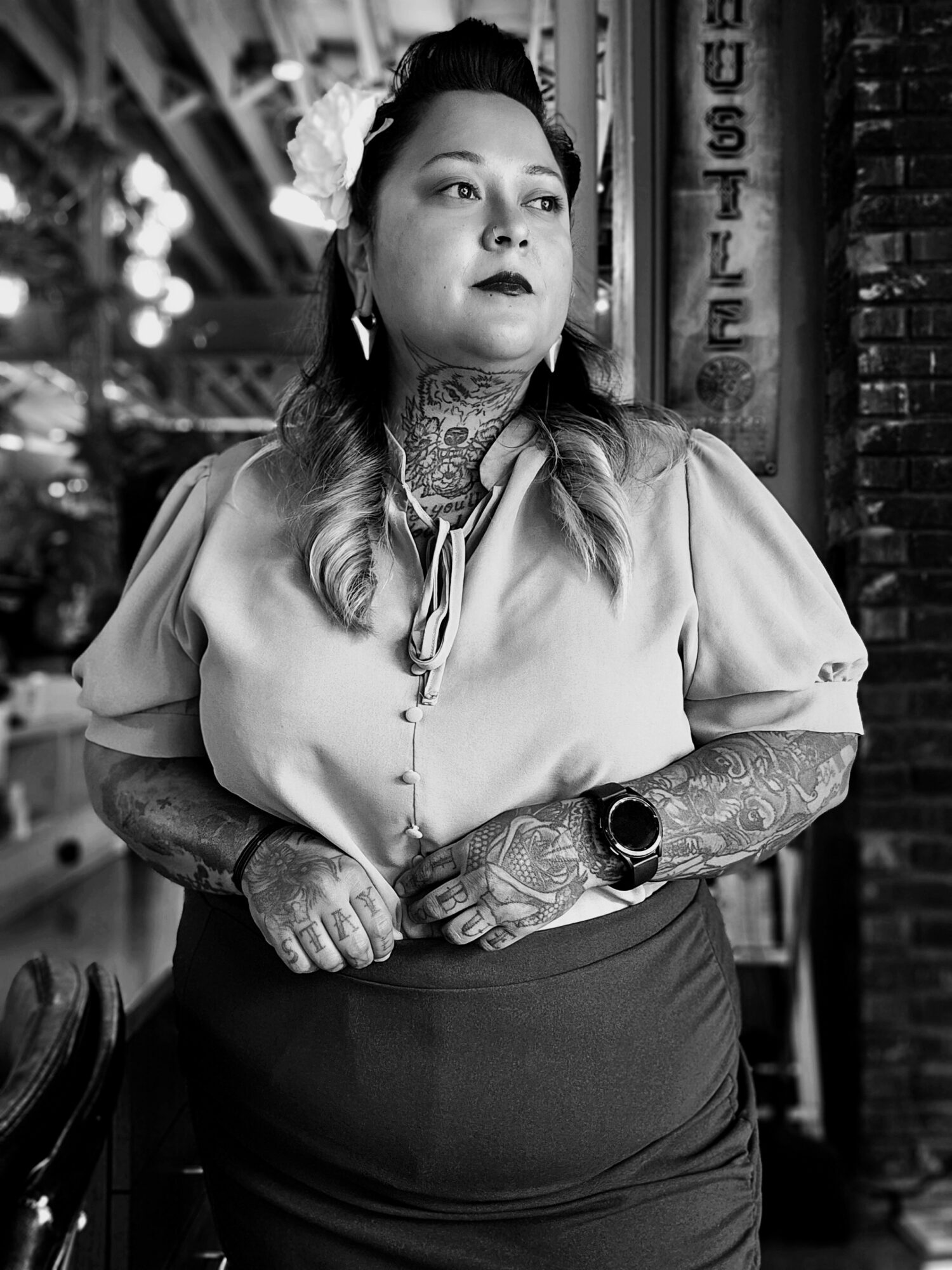 Woman with tattoos, wearing a blouse and skirt, standing outdoors near a brick wall and sign, looking to the side.